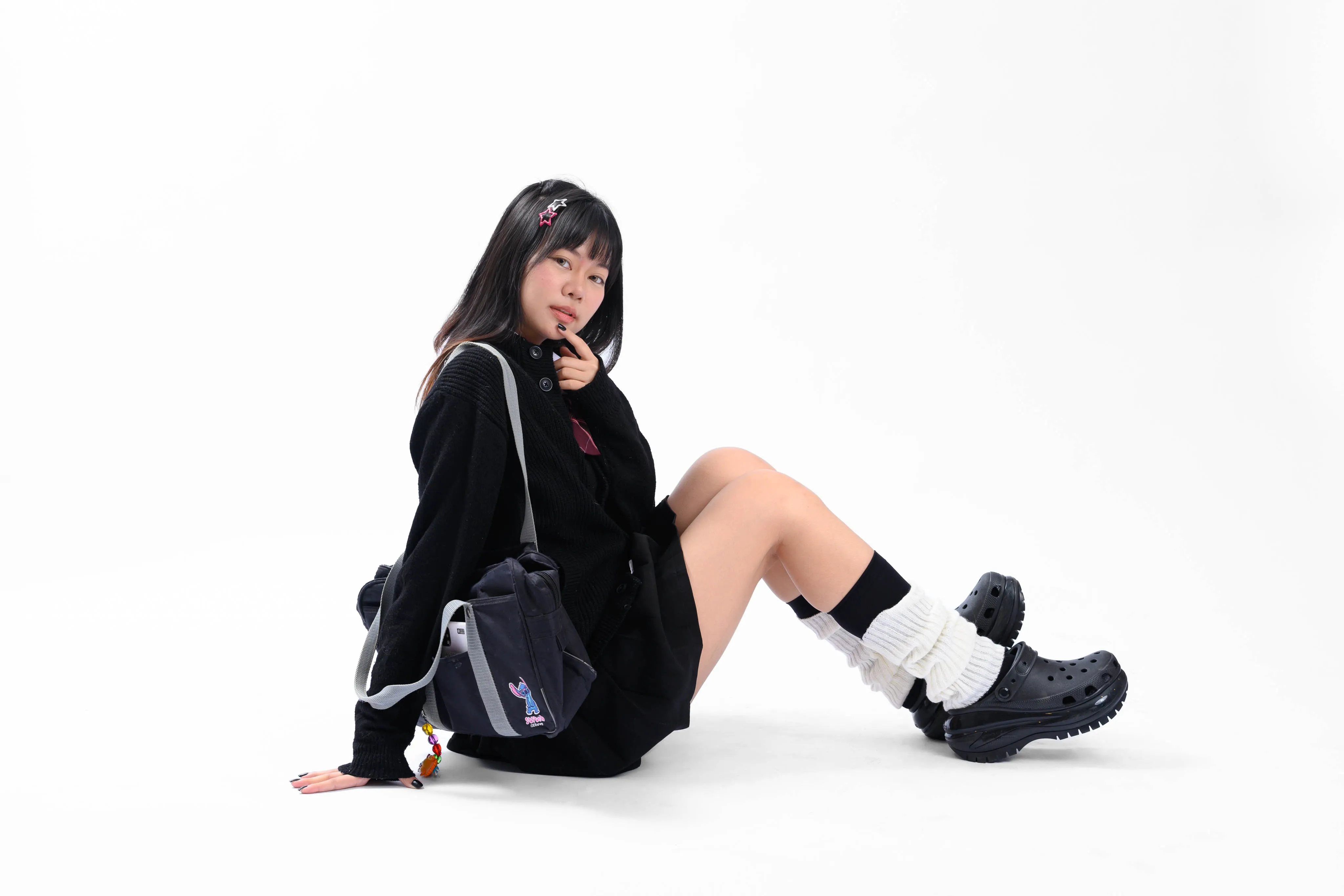 A young woman in a black uniform with a backpack and chunky clogs sitting on a white studio floor