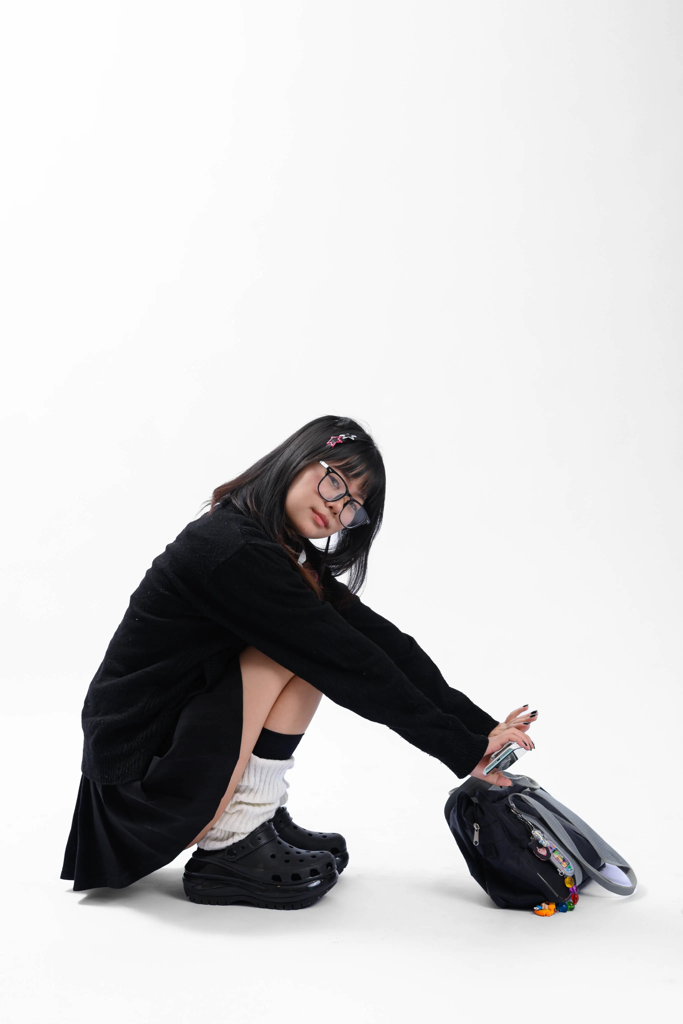 A young woman in glasses and a black uniform crouching with her backpack on a white studio backdrop