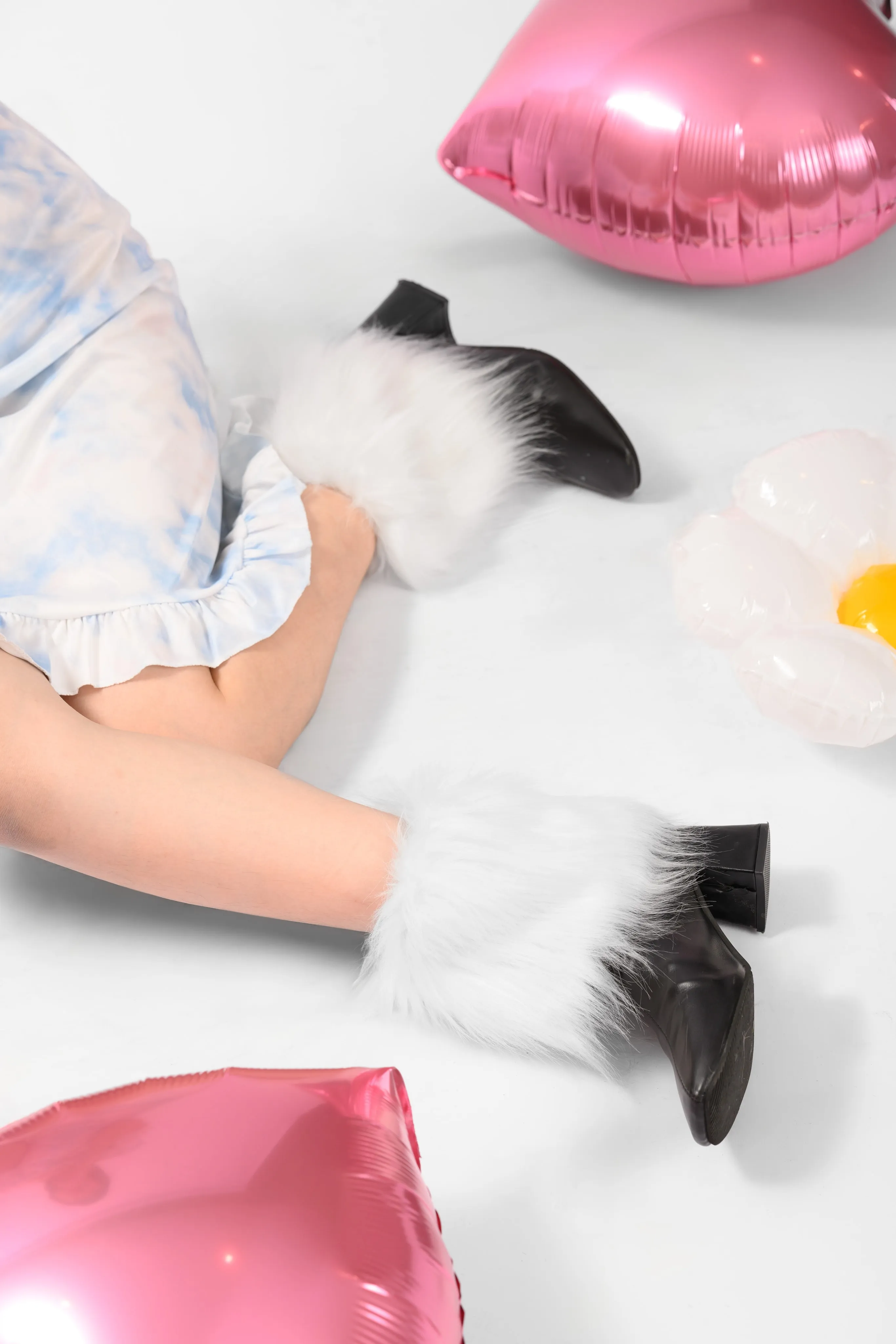 Legs in fur-trimmed black ankle boots and a pastel ruffled dress among pink heart balloons on white
