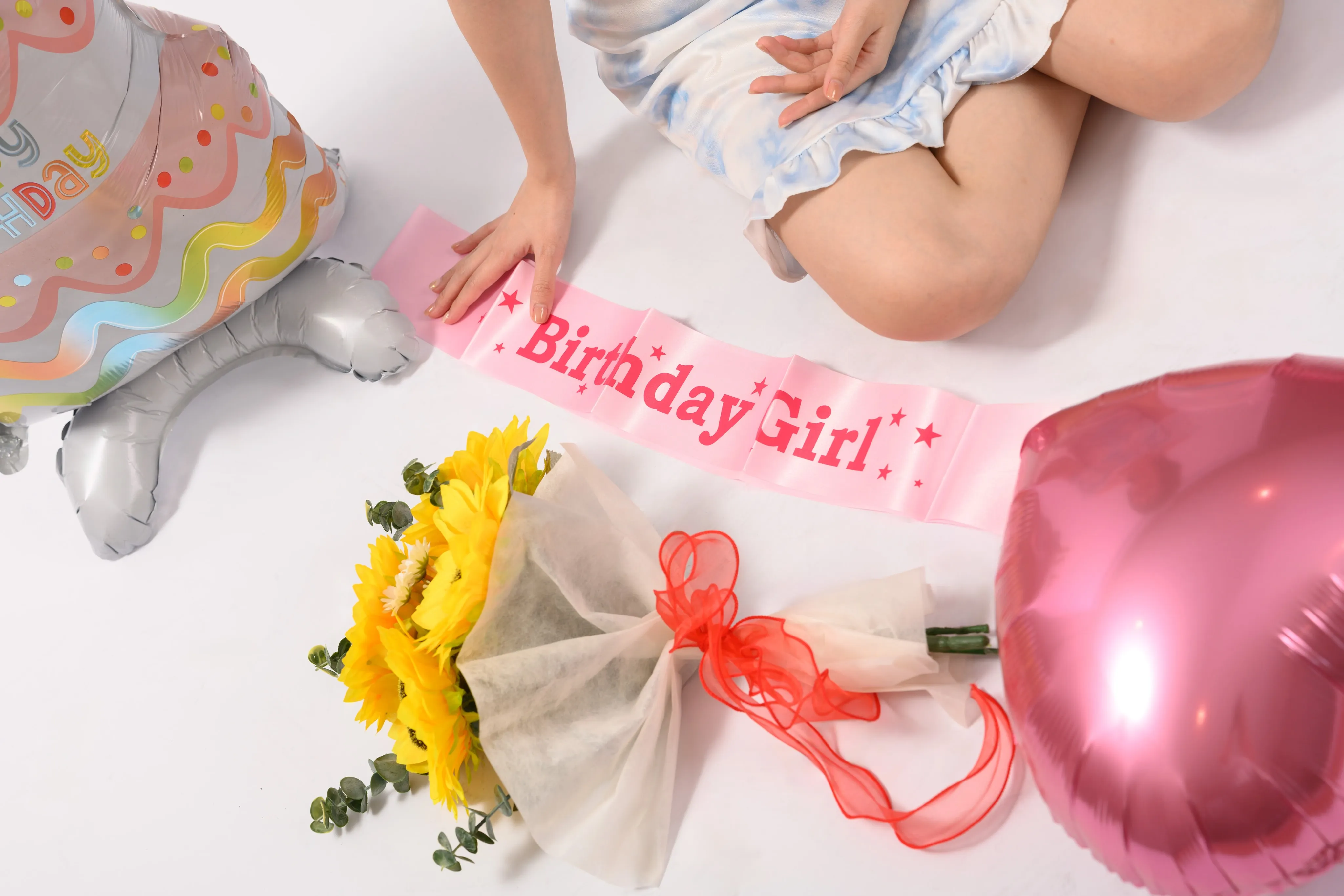 A "Birthday Girl" sash, sunflower bouquet, and pink balloons on a white studio floor beside a seated model