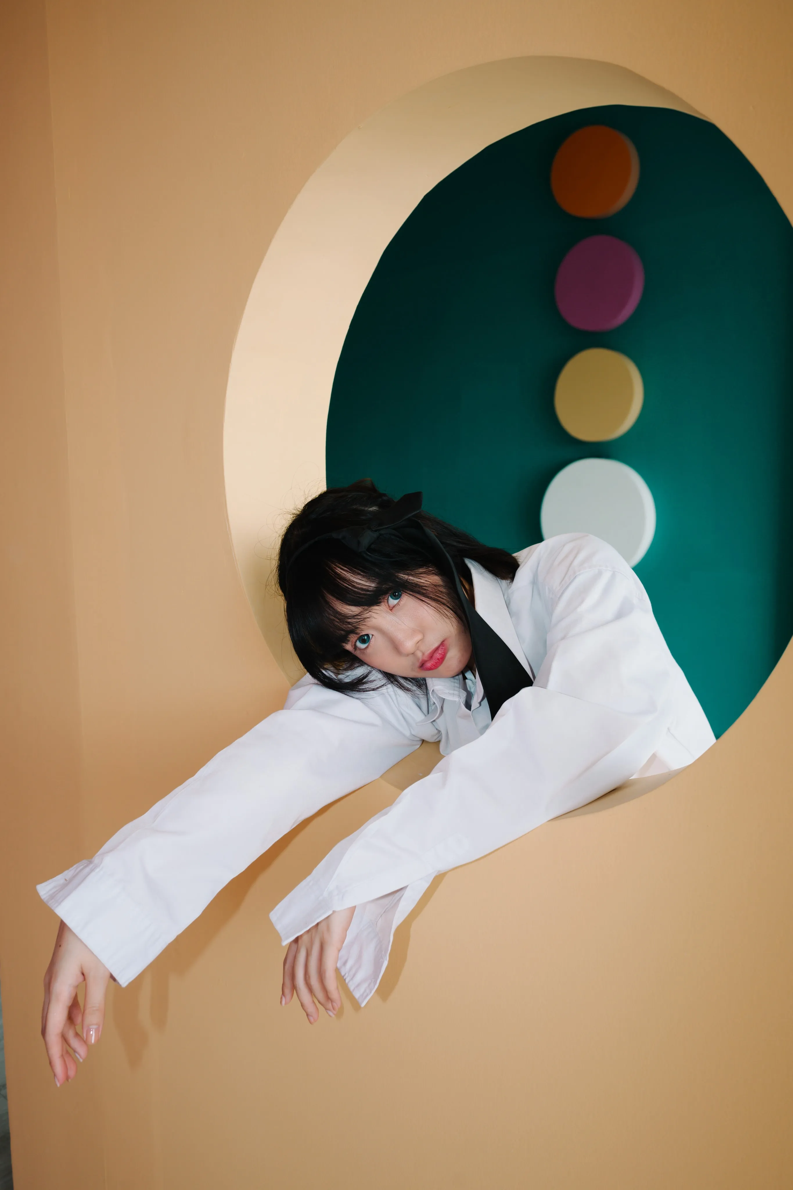 A young woman in a white blazer leaning out through a round opening in a peach-colored wall