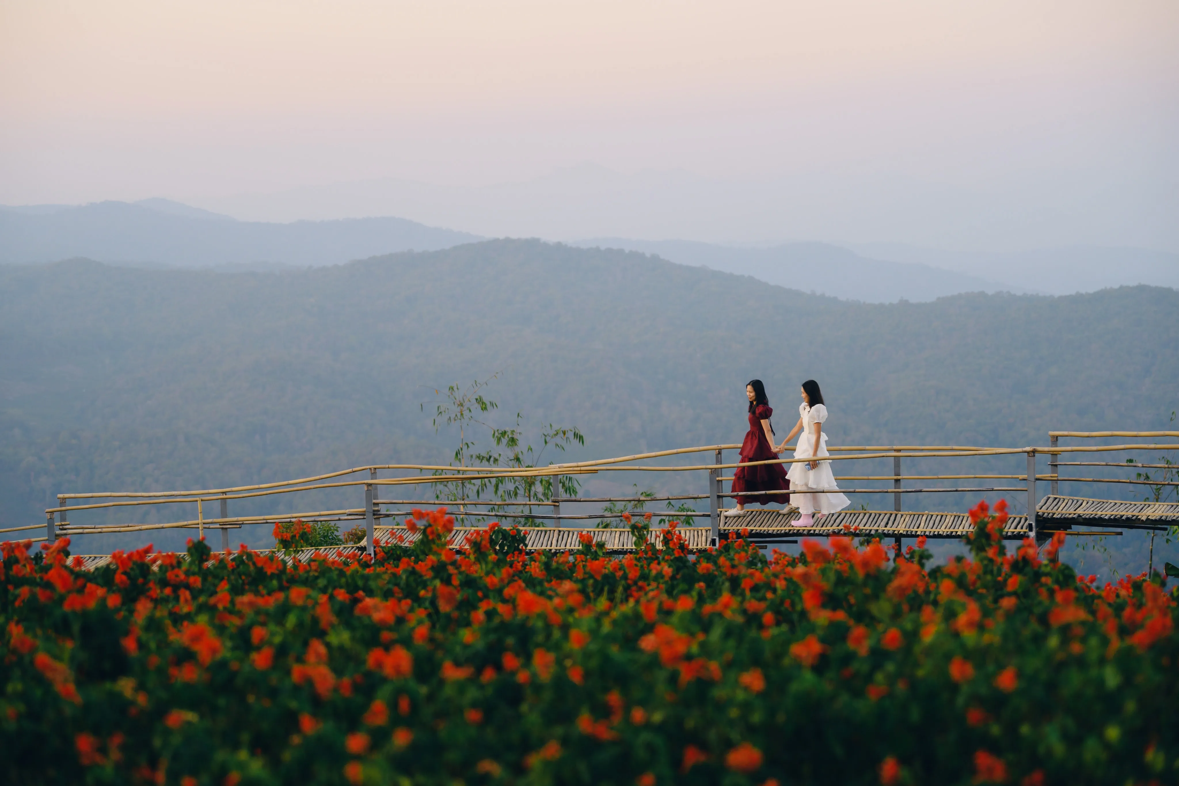 A couple in matching red and white dresses holding hands on a bamboo hillside walkway, mountains beyond.