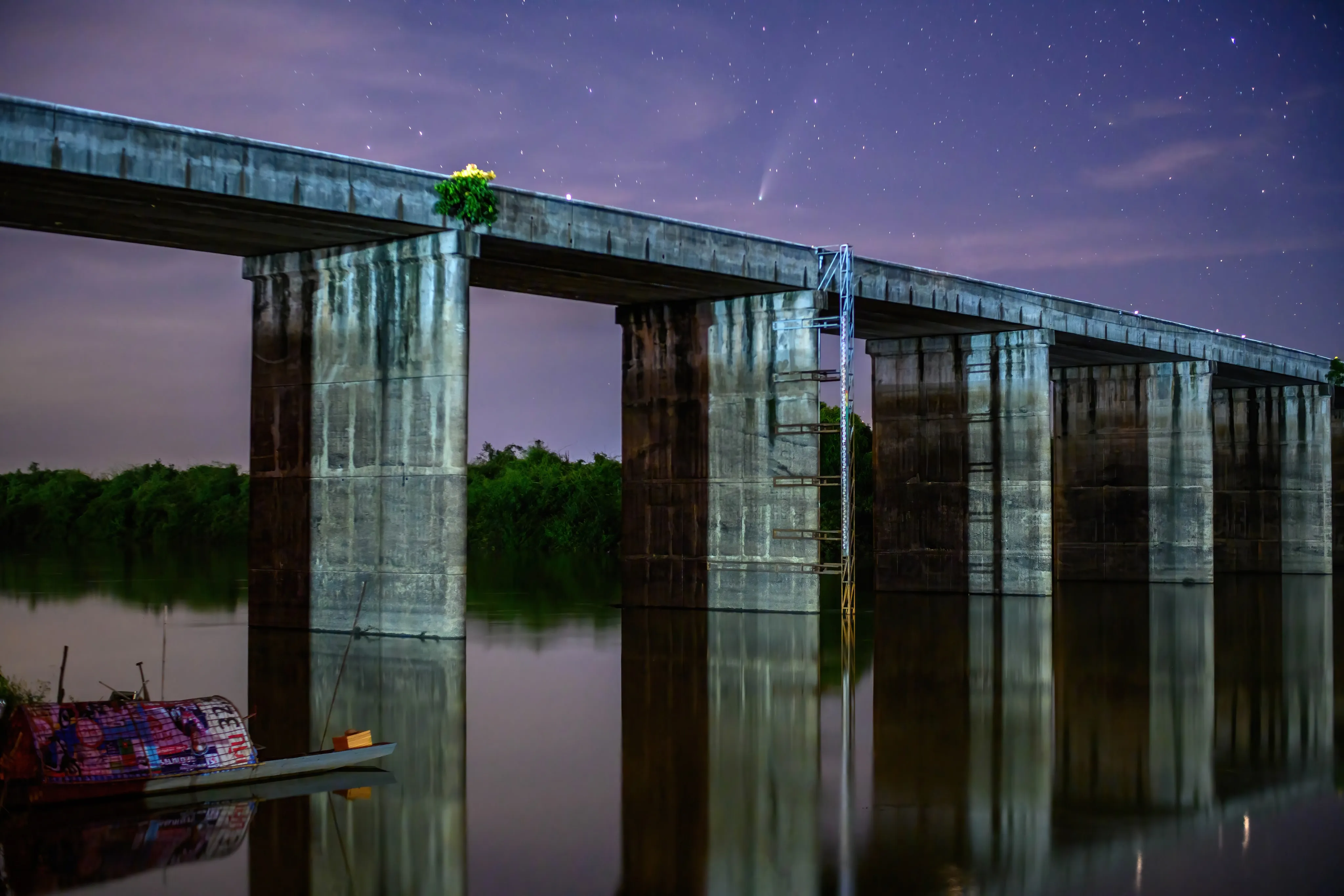 Comet NEOWISE streaking above a concrete railway bridge reflected in calm water