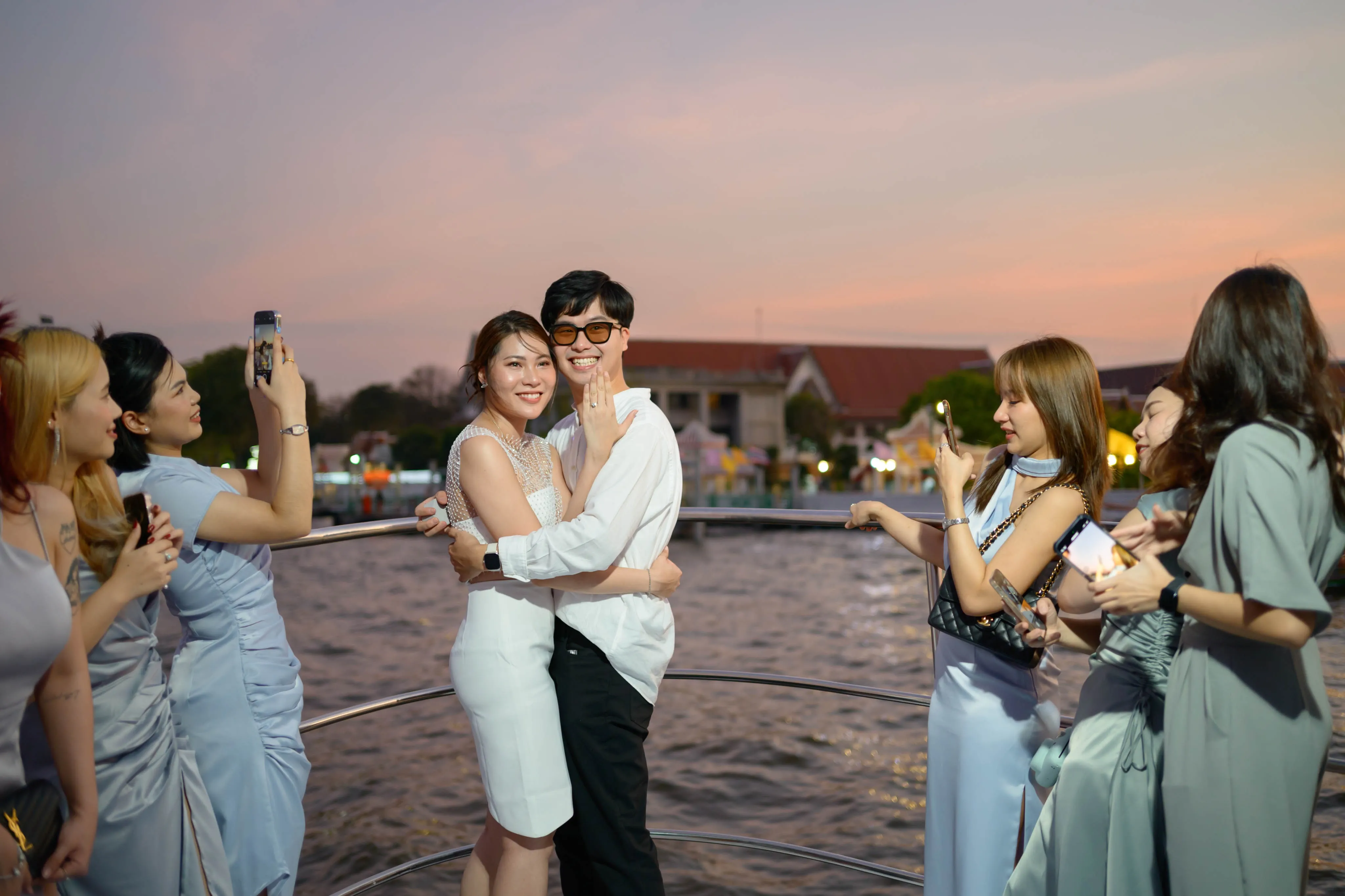 A couple embracing as friends celebrate at sunset beside the water