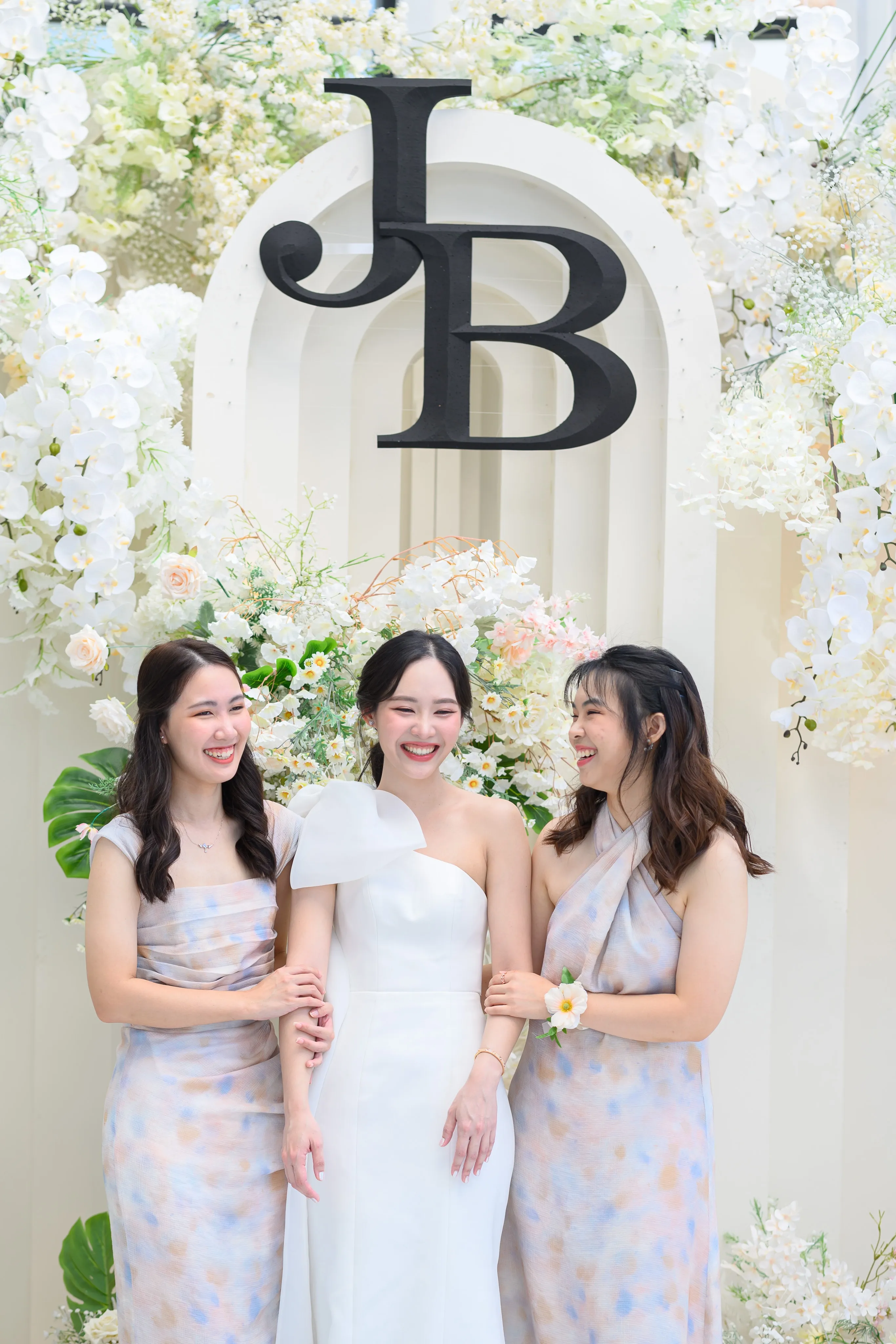 A smiling bride between two bridesmaids beneath a floral monogram arch