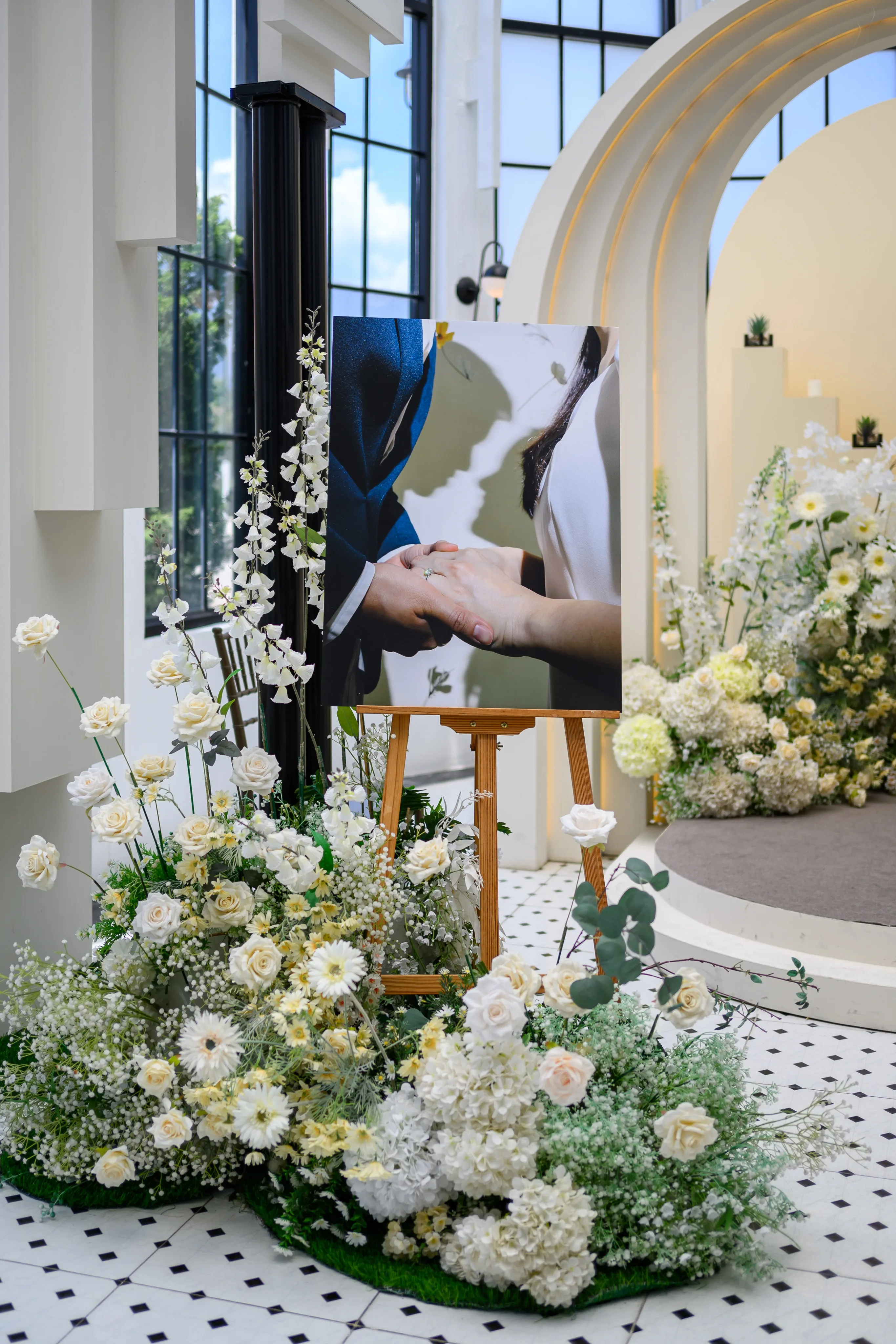 A framed photograph of the couple's joined hands on an easel amid white wedding flowers