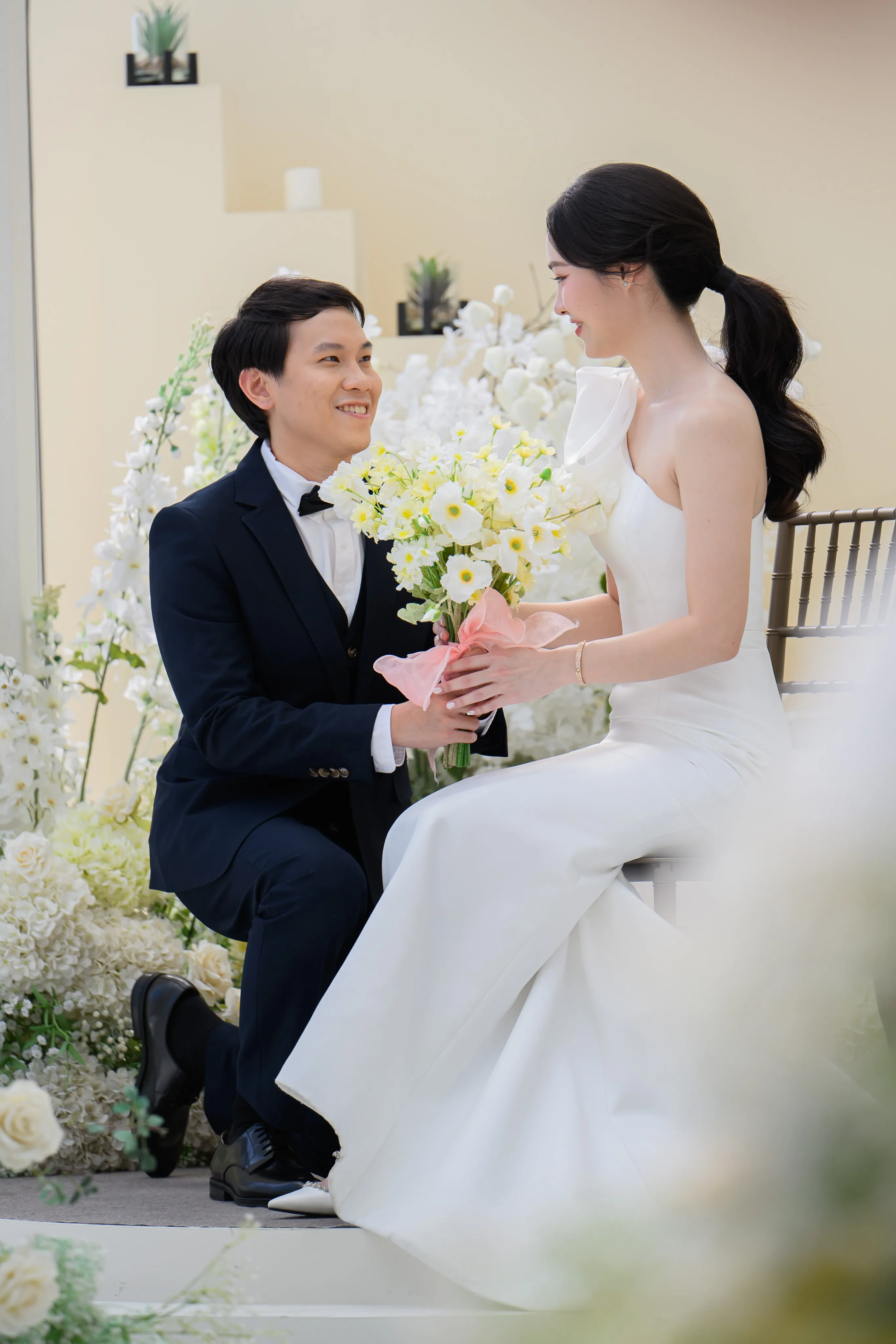 A groom kneeling to offer a bouquet to the seated bride in a flower-filled room