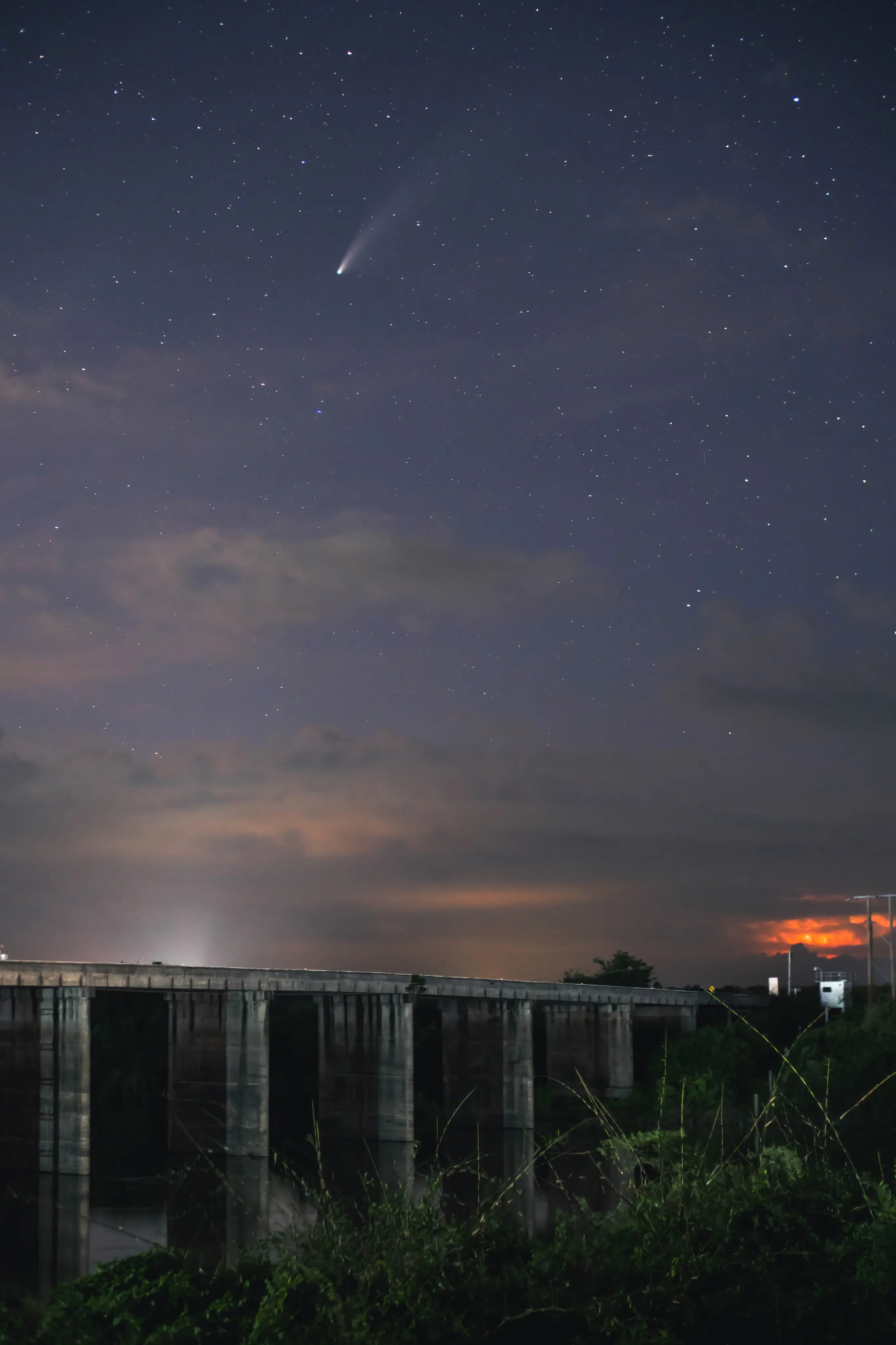 Comet NEOWISE above a dam, with orange lights glowing on the horizon