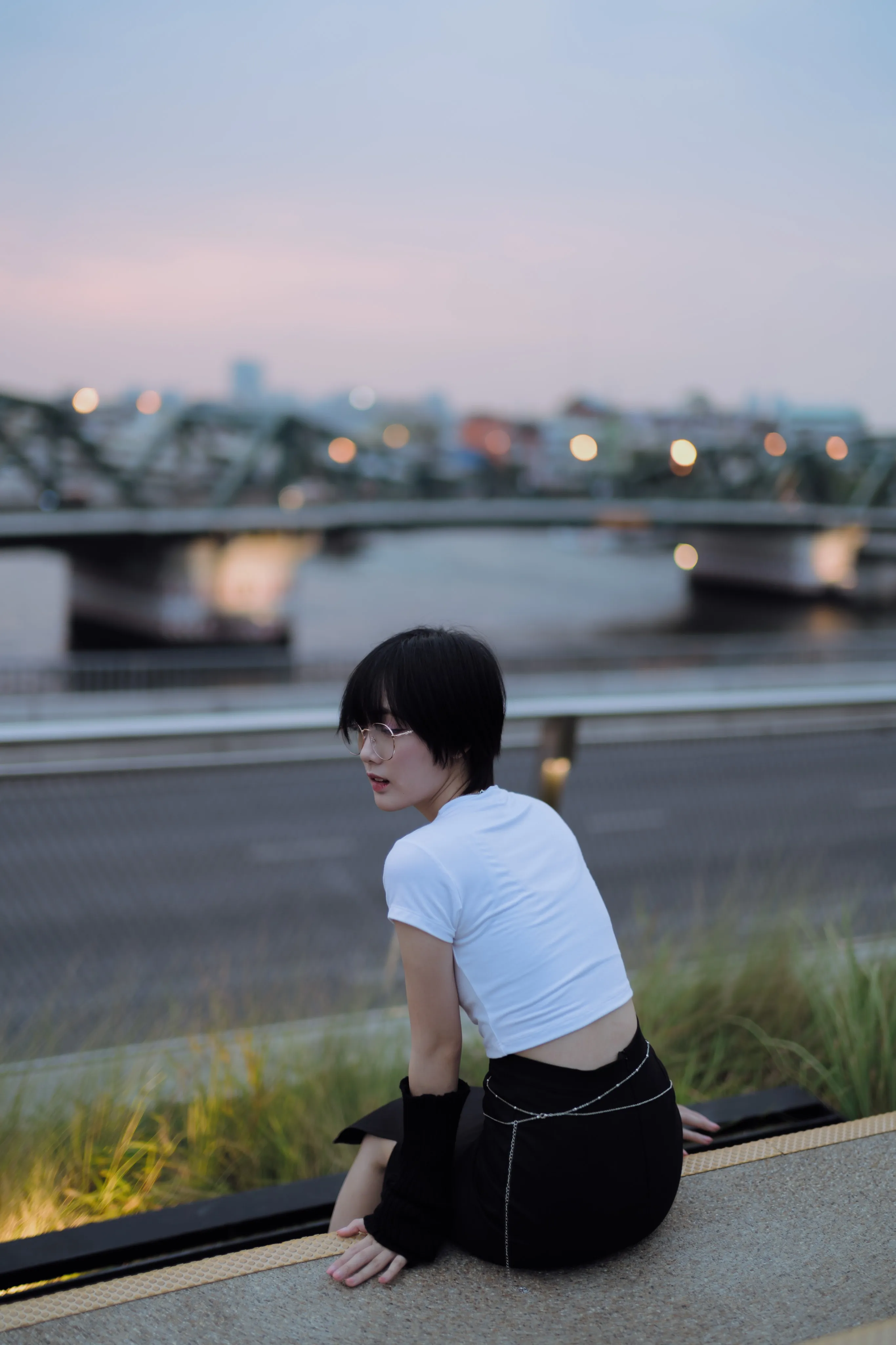 A woman seated beside a road at dusk with a bridge in the distance