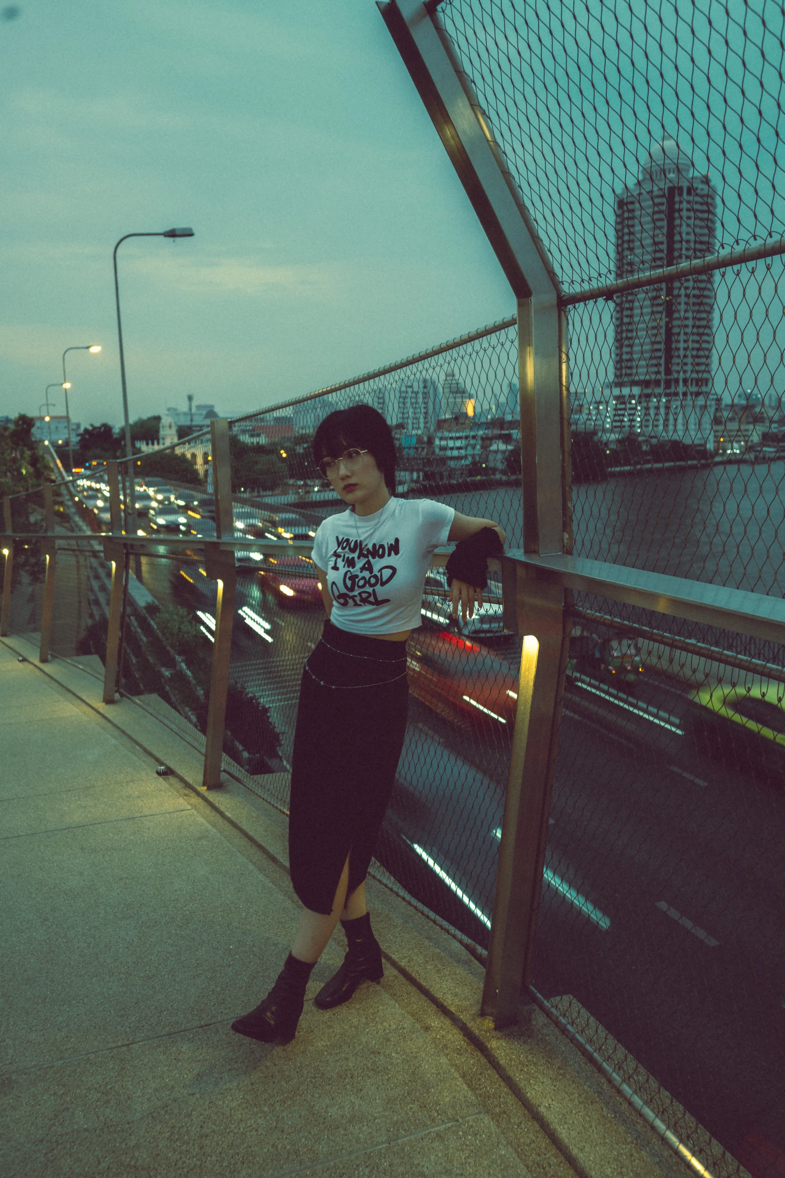 A woman posing beside a chain-link fence above a busy road