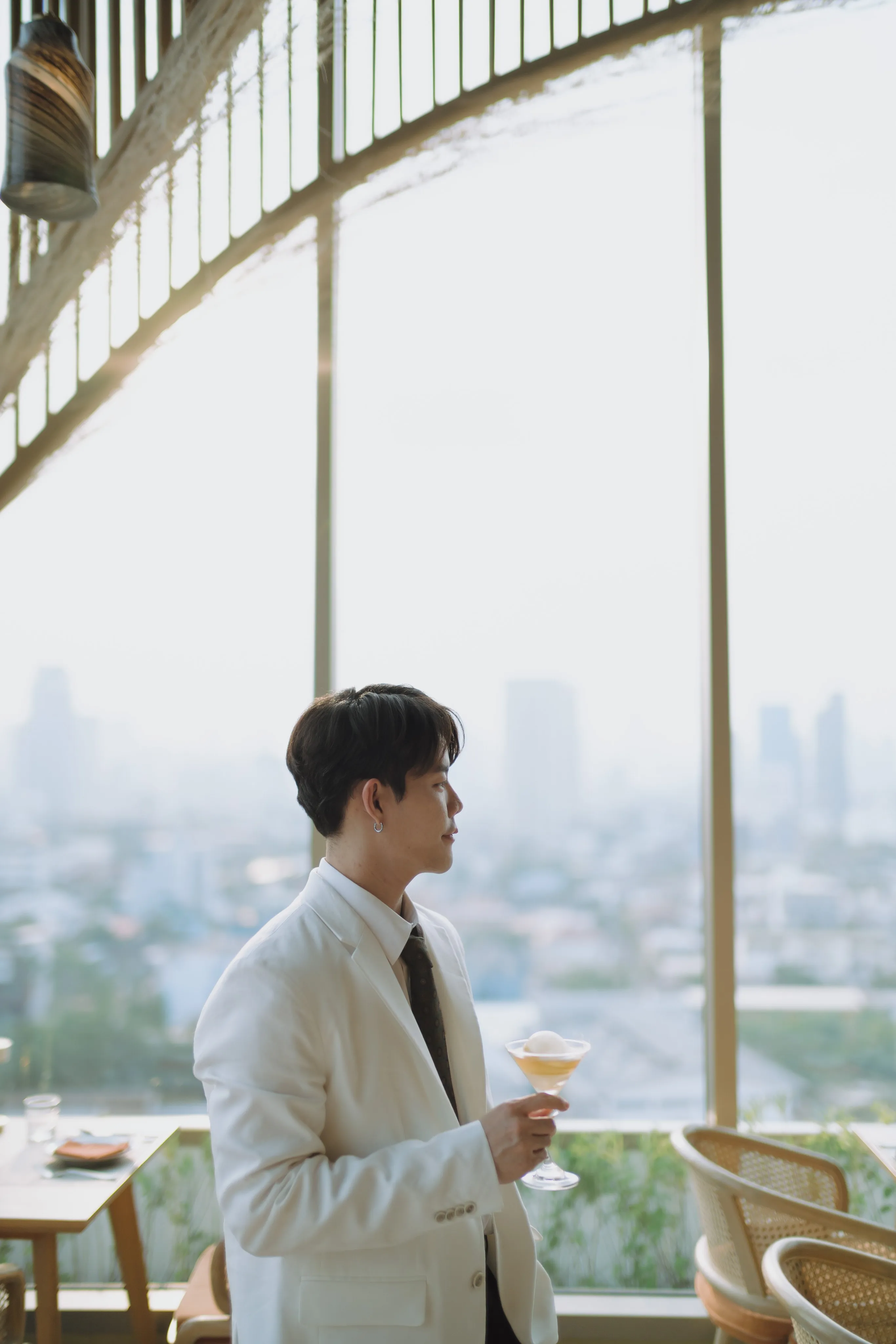 A man in a light jacket holding a drink beside a bright window