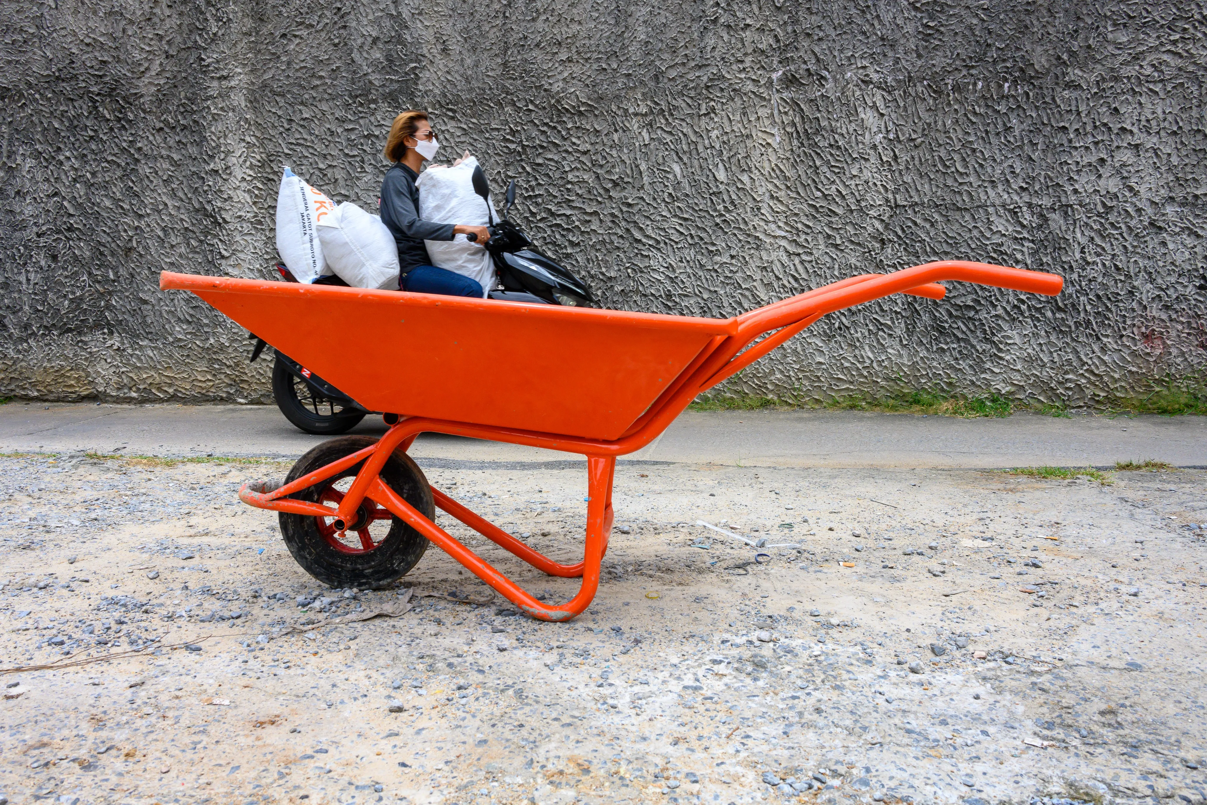 An orange wheelbarrow on gravel as a masked motorbike rider carrying sacks passes a gray wall