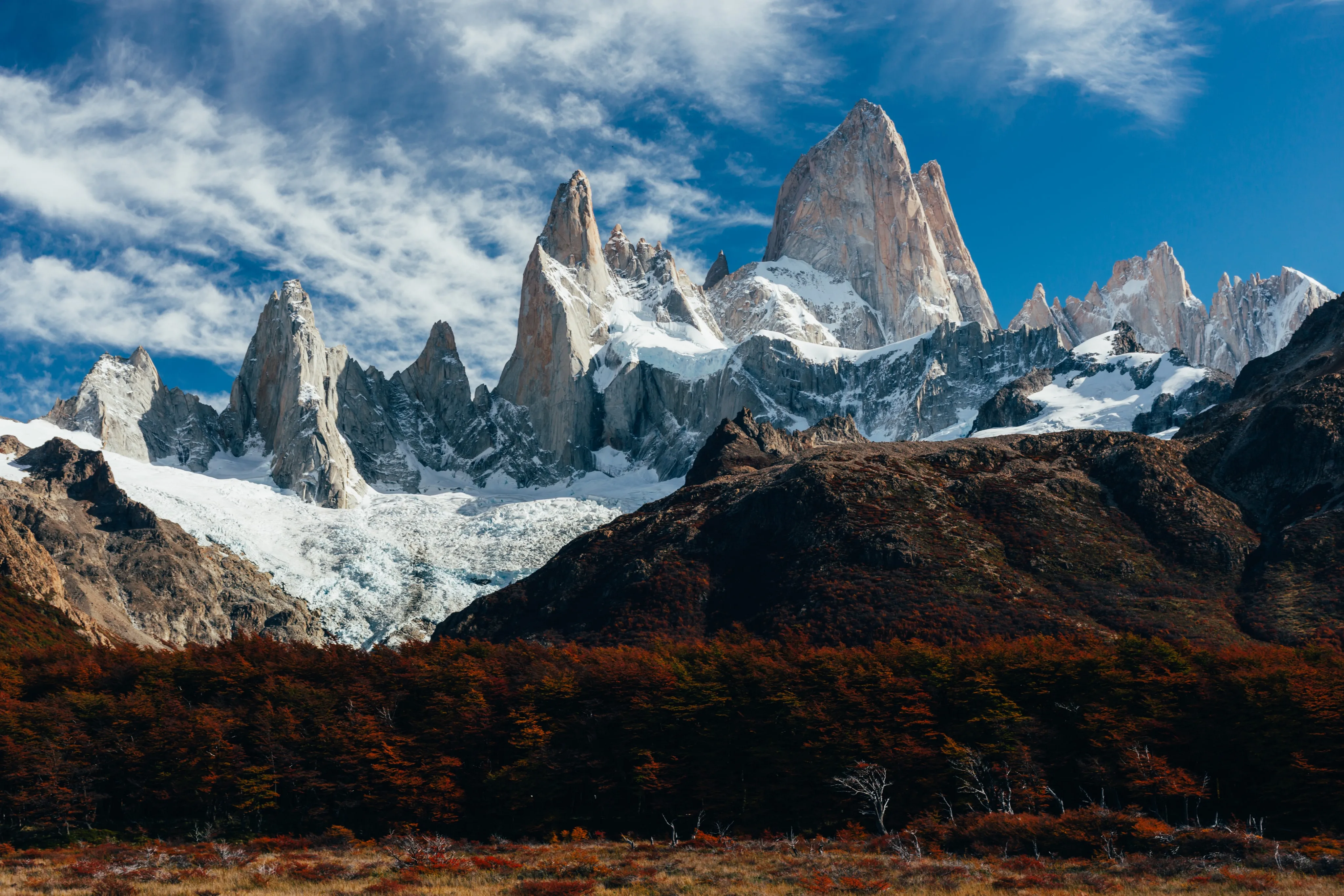 Jagged granite spires above a glacier and autumn-colored forest under a cloud-streaked sky