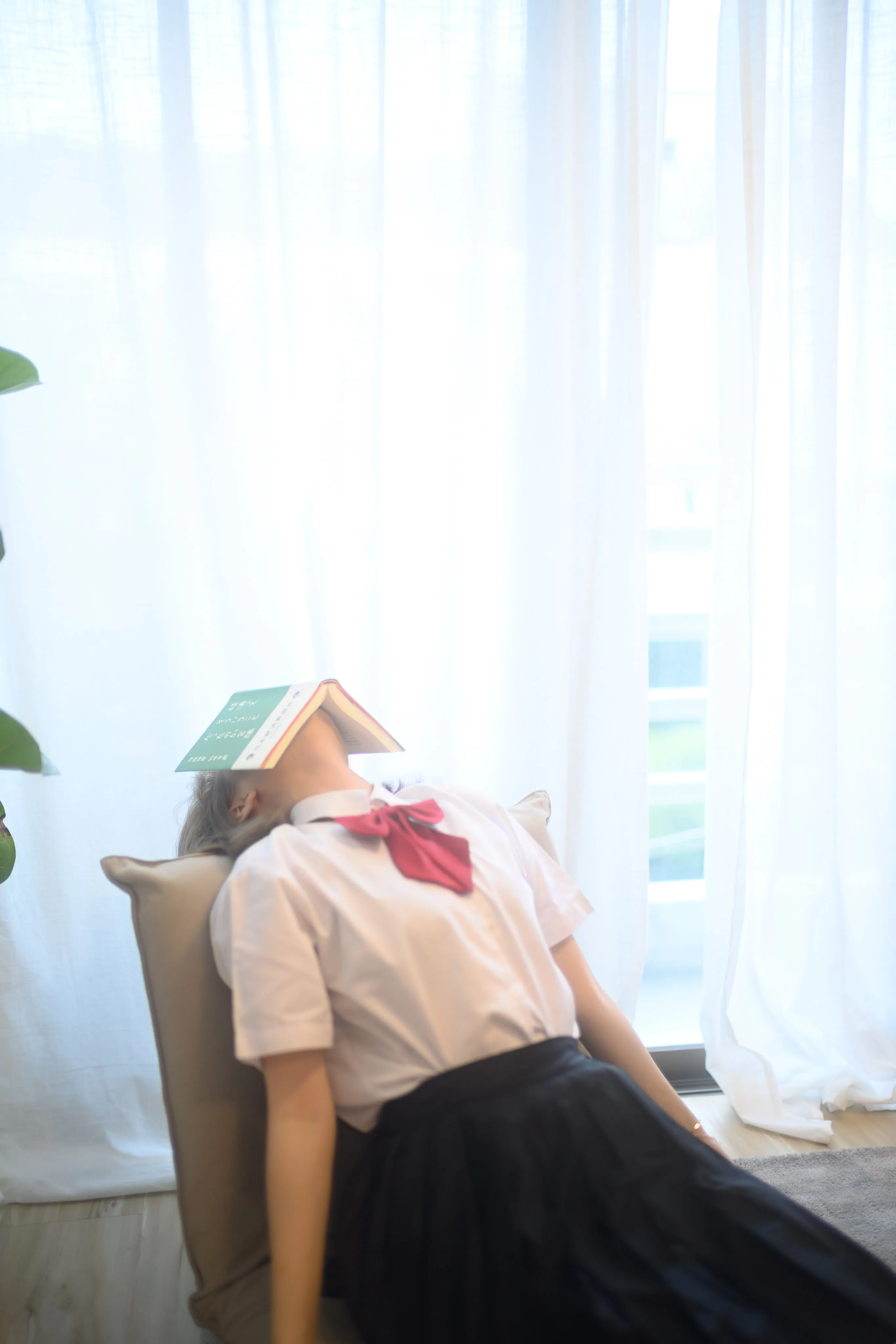 A young woman in a school uniform reclining in a chair beside a bright window, an open book covering her face
