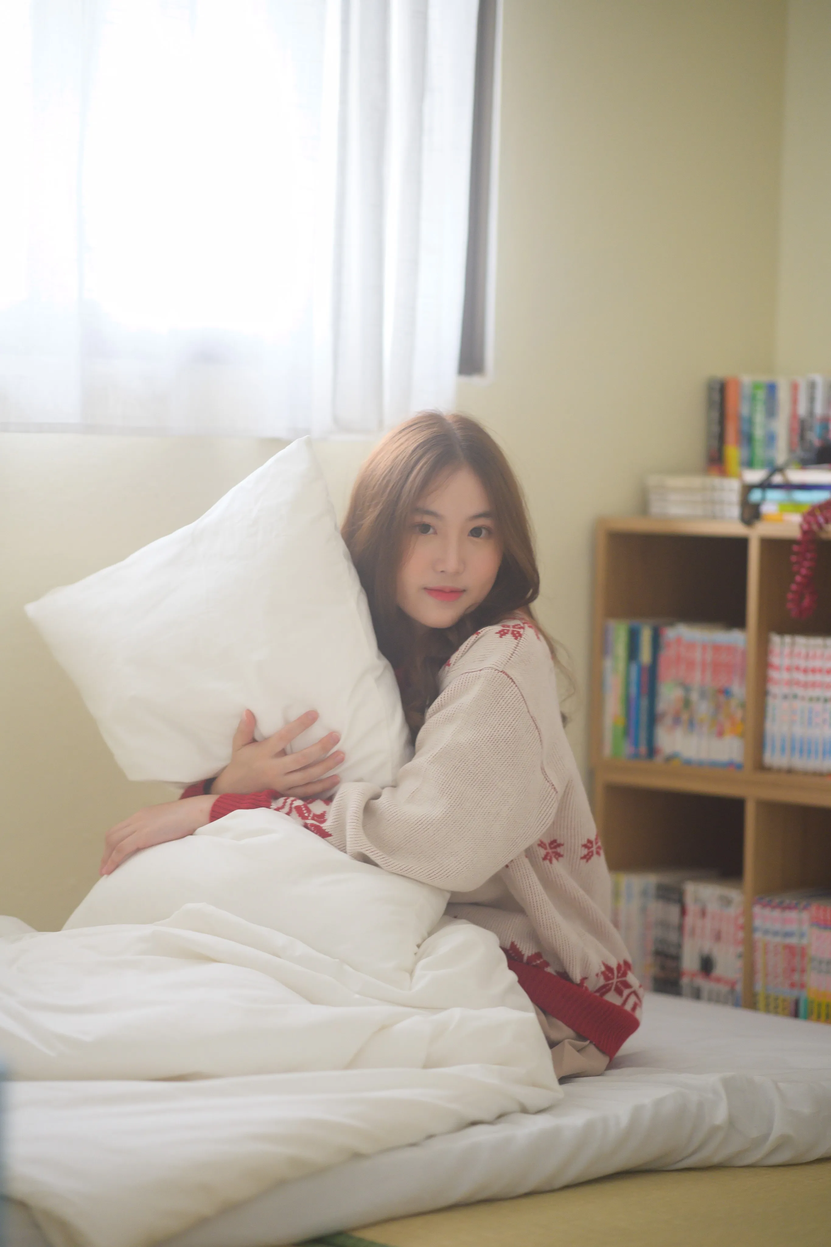 Woman hugging a white pillow while sitting on a bed beside a bookshelf