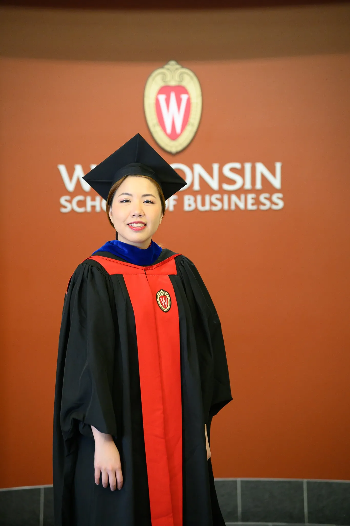 Graduate in black and red regalia posing beneath a Wisconsin School of Business emblem