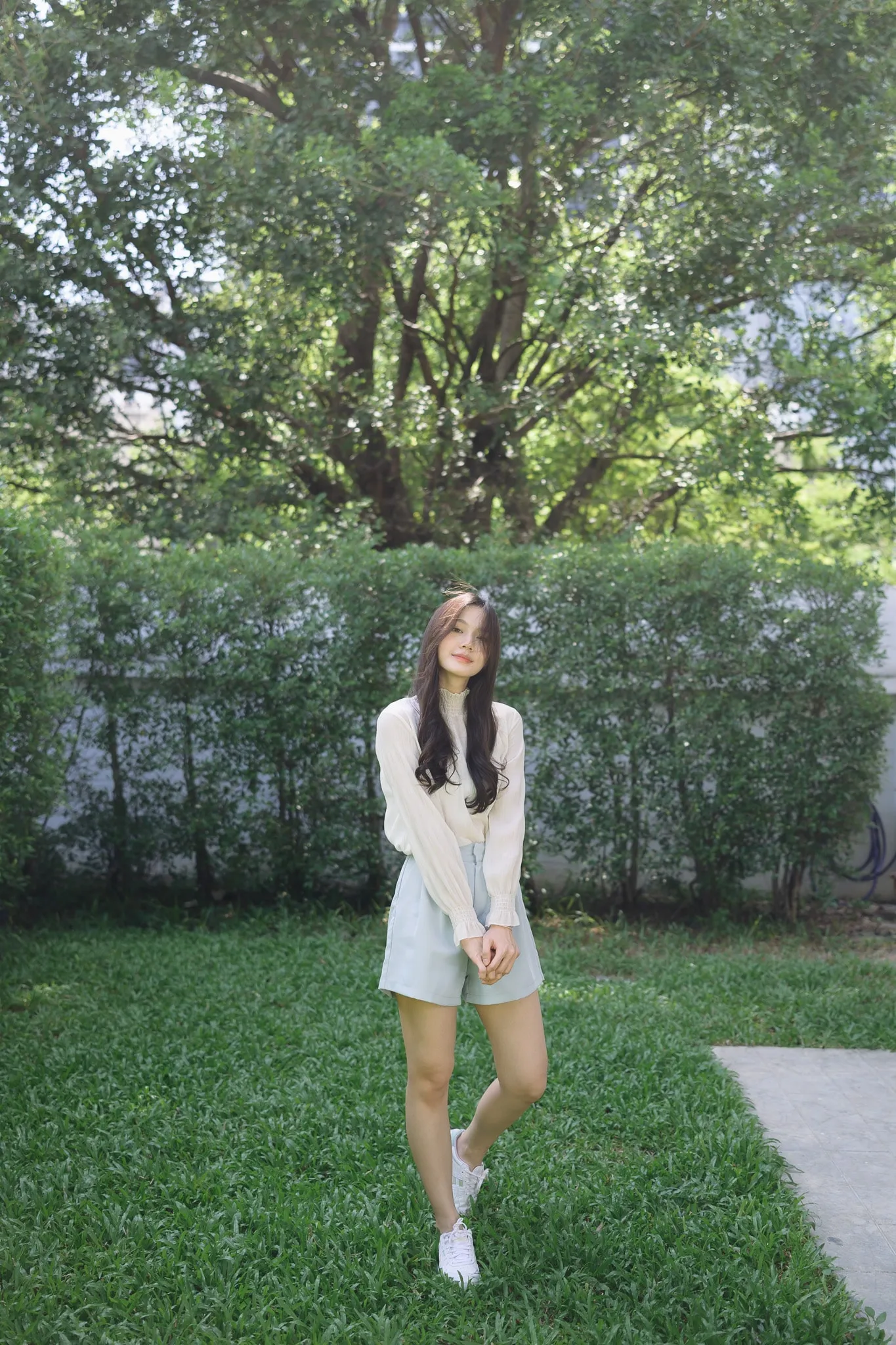 A woman in a cream cardigan and light shorts standing on a lawn in front of a hedge and large trees
