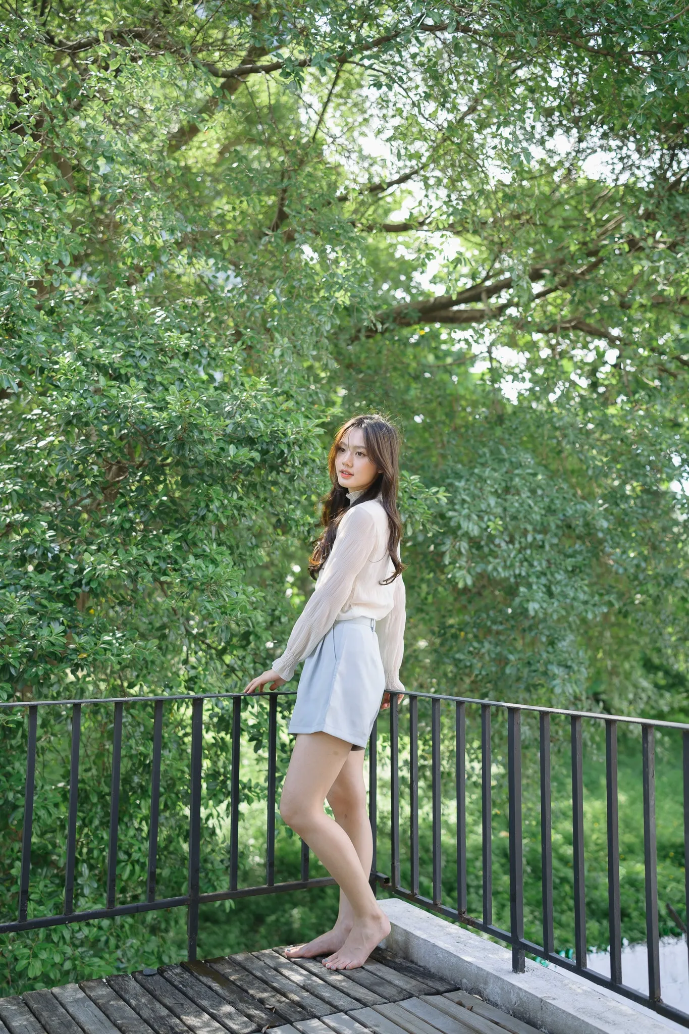 A barefoot woman in a cream cardigan leaning on a black railing on a wooden deck surrounded by trees