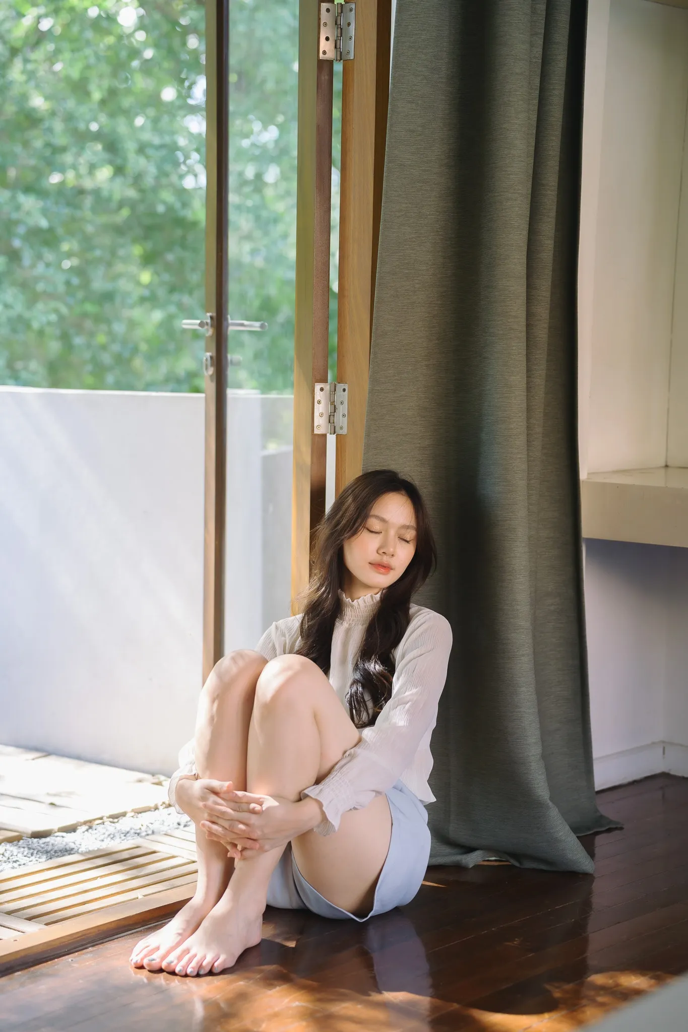 A barefoot woman hugging her knees on a sunlit wooden floor beside an open glass door and a long curtain