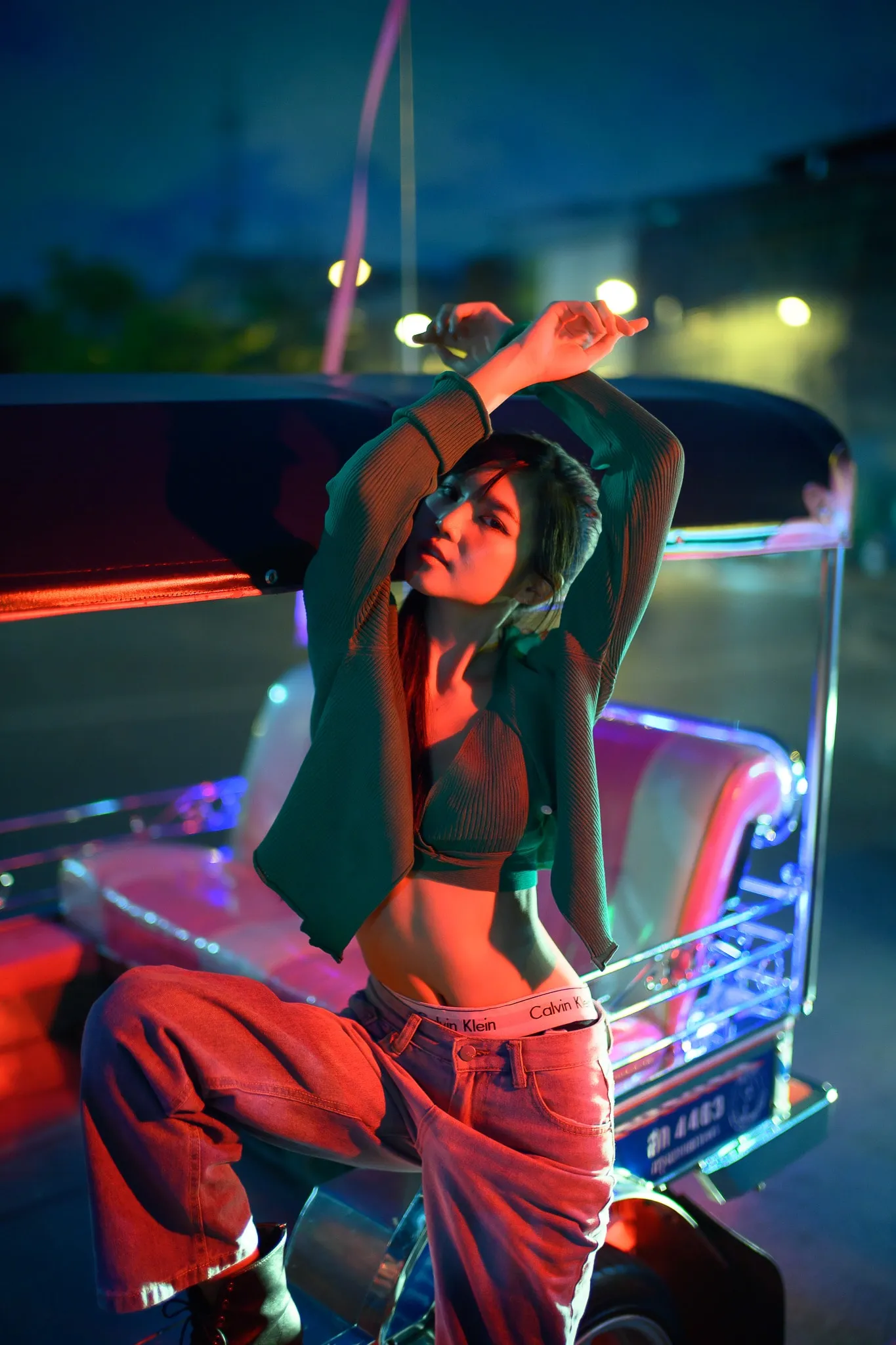A woman in a cropped top and baggy red trousers posing with her arms raised beside a neon-lit tuk-tuk at night