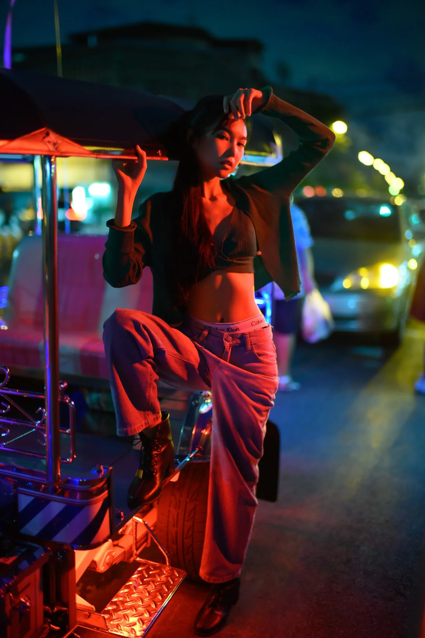 A woman in a cropped jacket and red trousers standing on the step of a neon-lit tuk-tuk with one arm raised