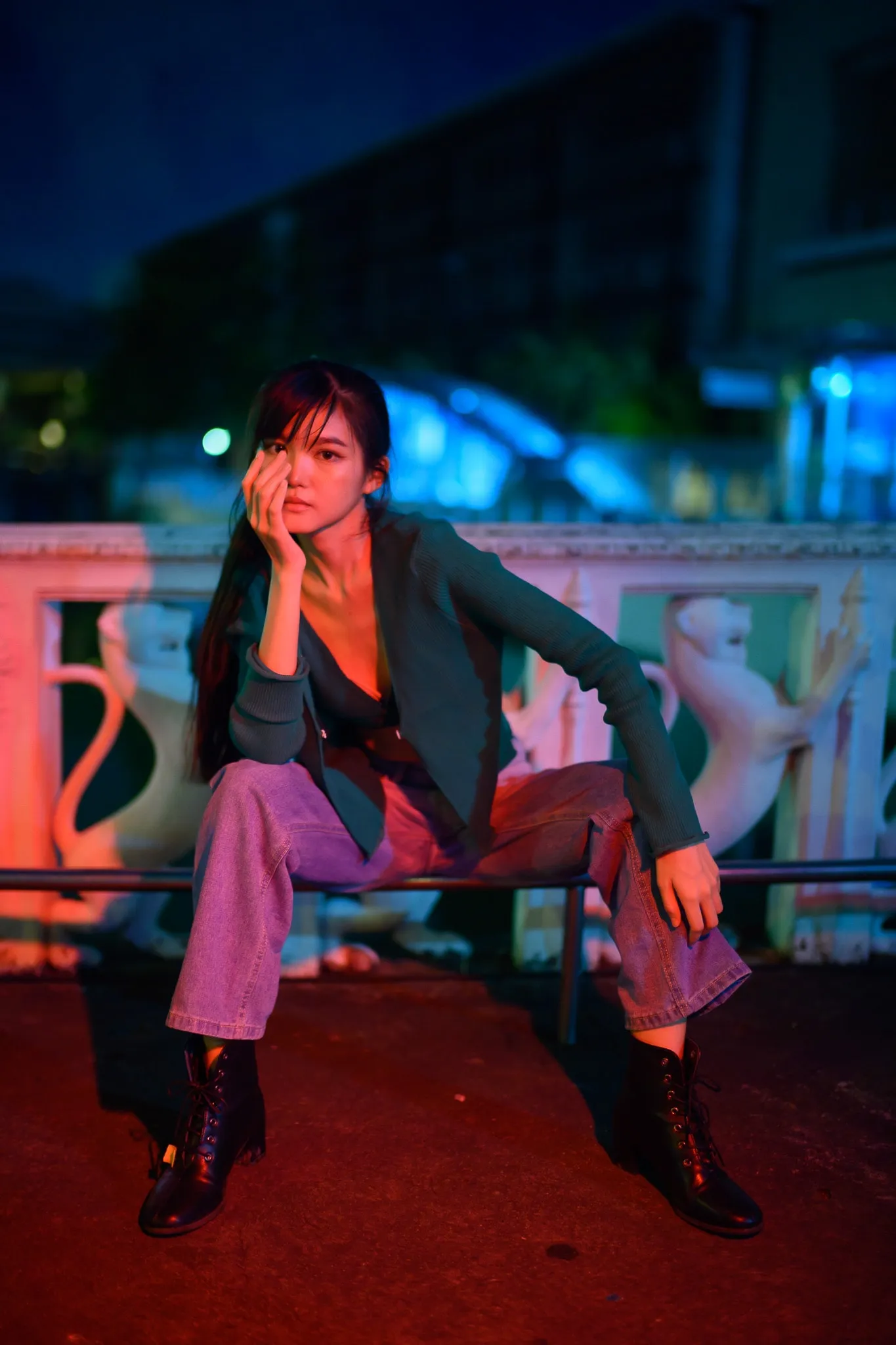 A woman in a dark blazer and jeans sitting on a railing in vivid red and blue light at night