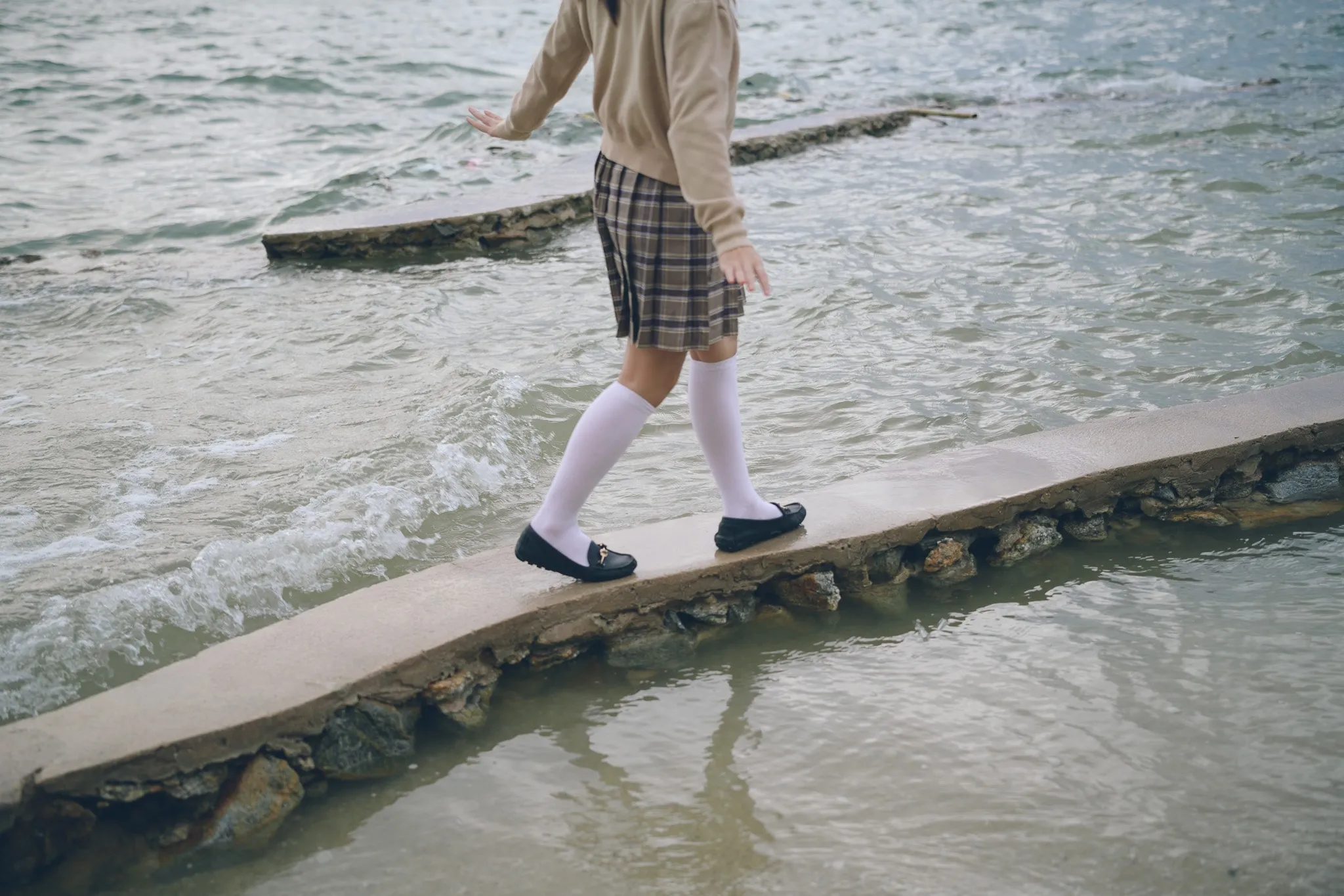 A woman in a plaid skirt and white knee socks walking along a narrow concrete ledge through shallow surf