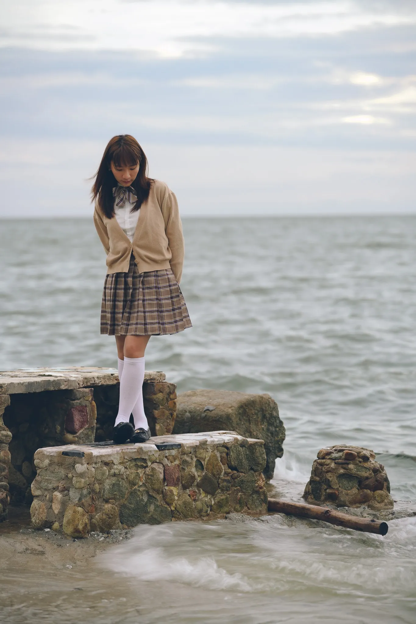 A woman in a beige cardigan and plaid skirt standing on rocks beside a grey sea
