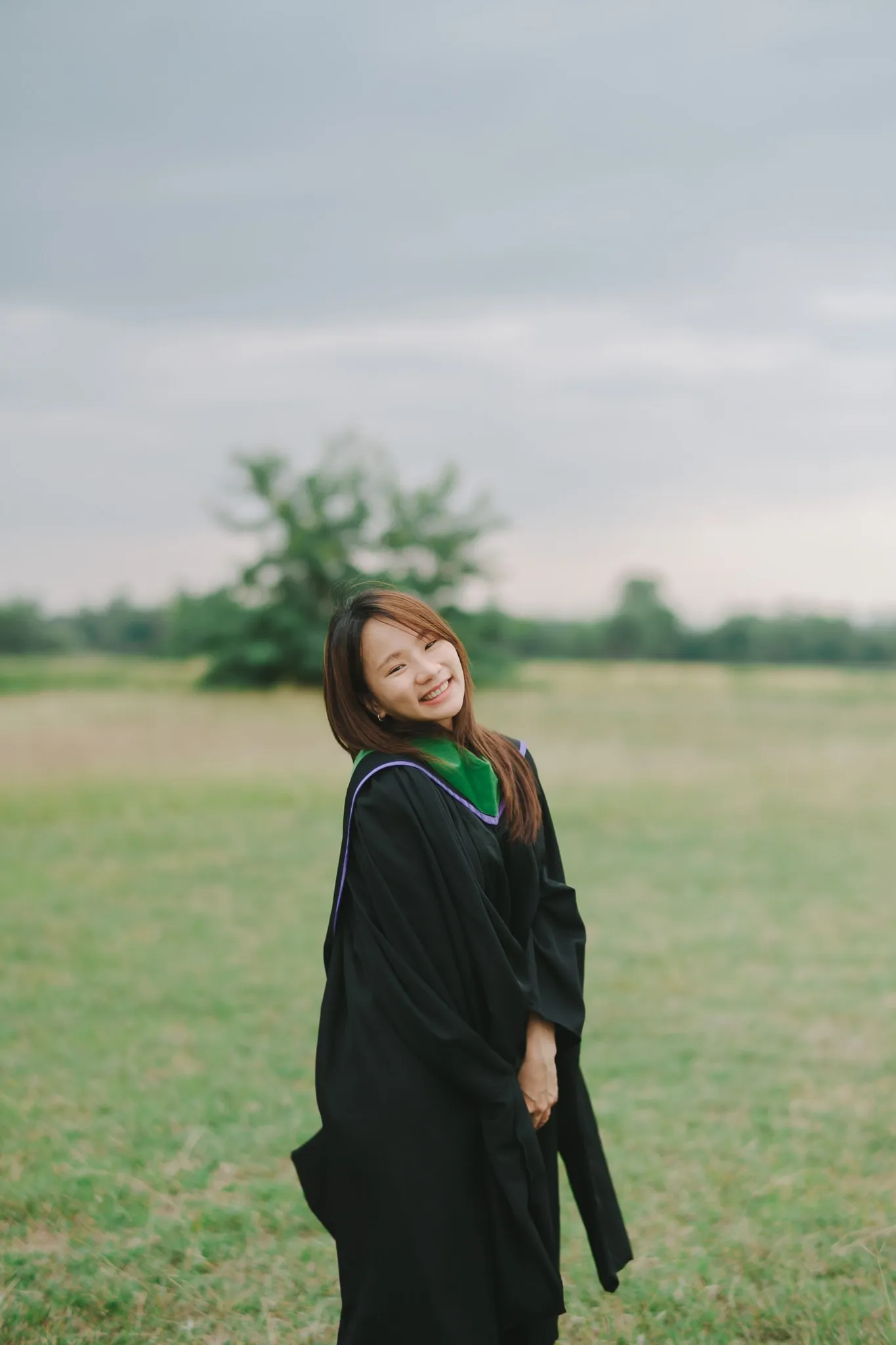 A smiling graduate in a black gown with a green hood tilting her head in an open field