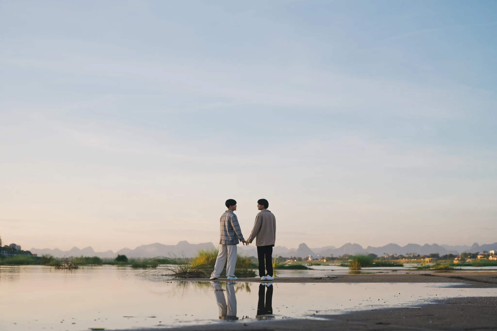Two people holding hands at the water's edge at dusk, reflected in the still shallows