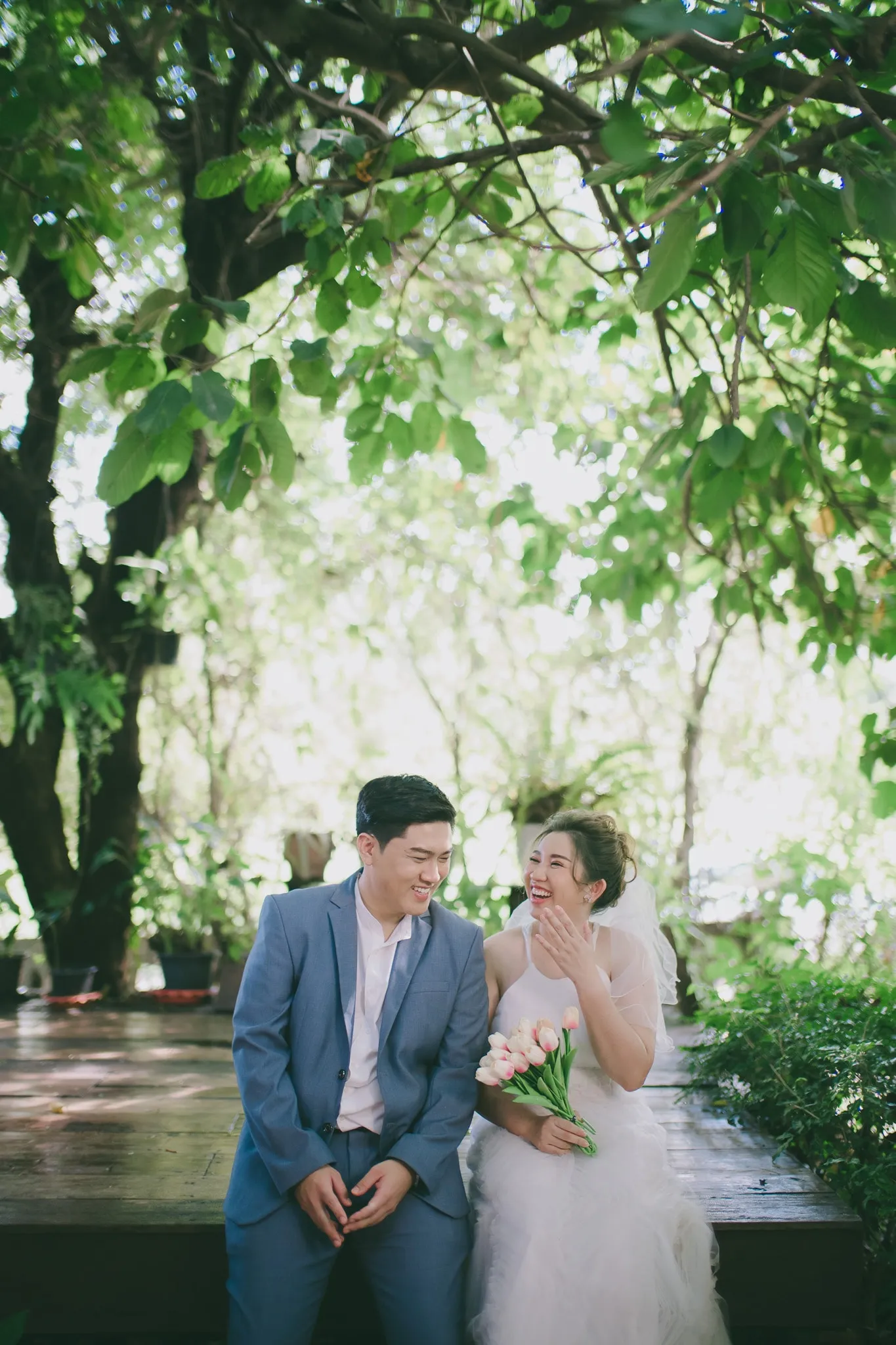 A laughing couple in a blue suit and white dress sitting together under leafy trees