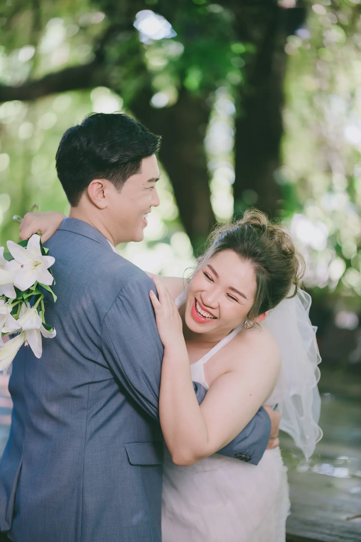 A laughing bride in a veil hugging her partner in a blue suit, holding white lilies