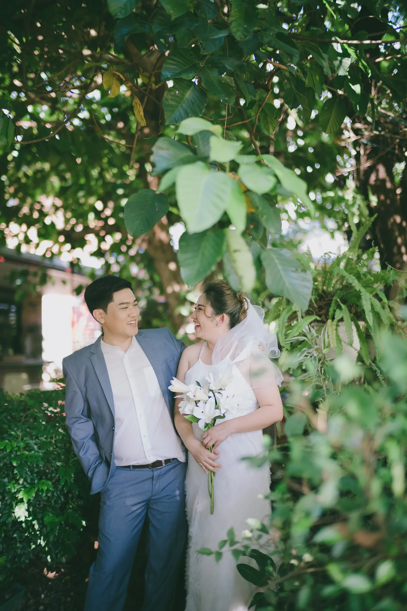 A couple in a blue suit and white dress standing arm in arm among green leaves