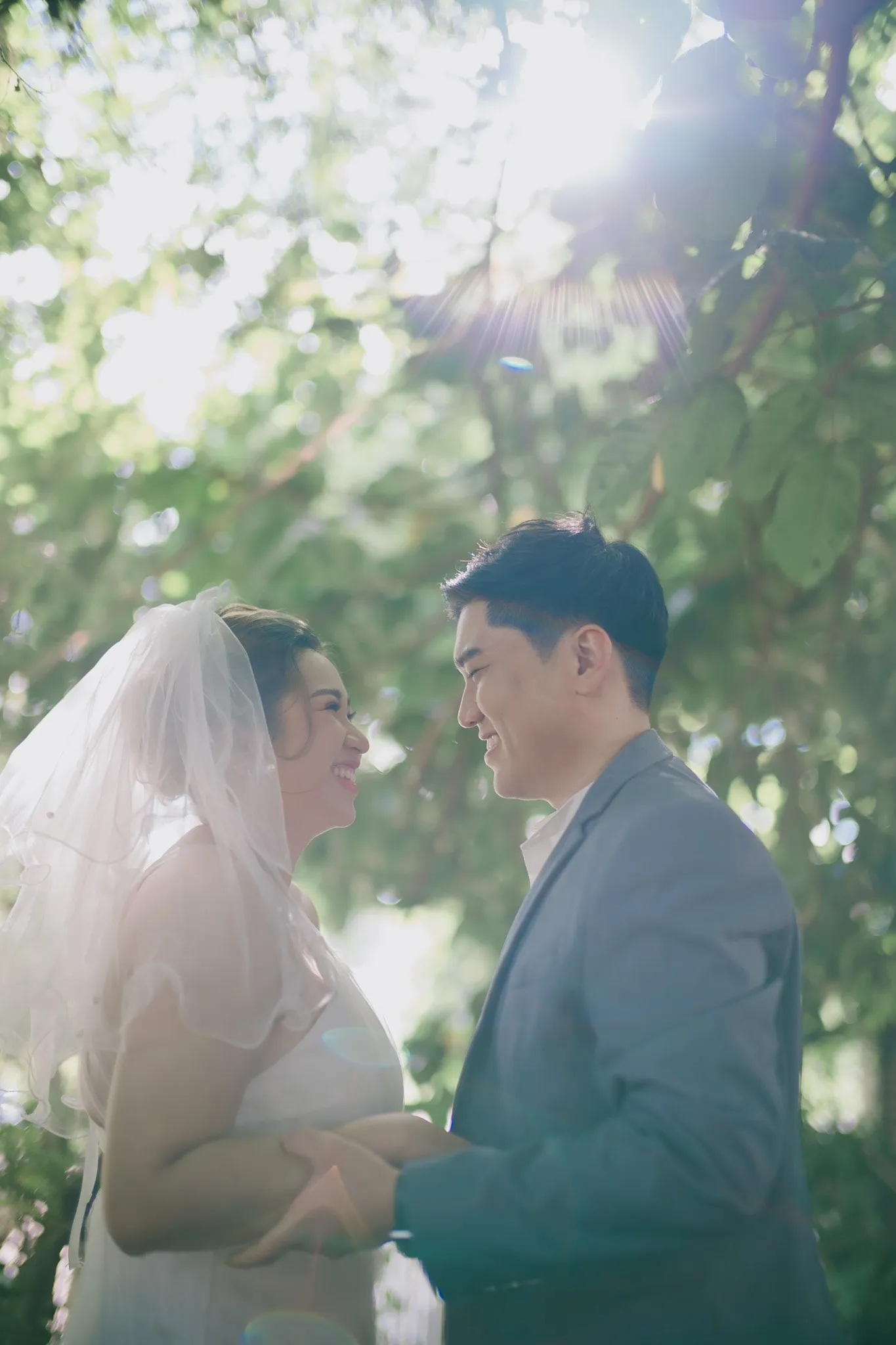 A bride in a veil and her partner in a grey suit holding hands in soft backlit greenery