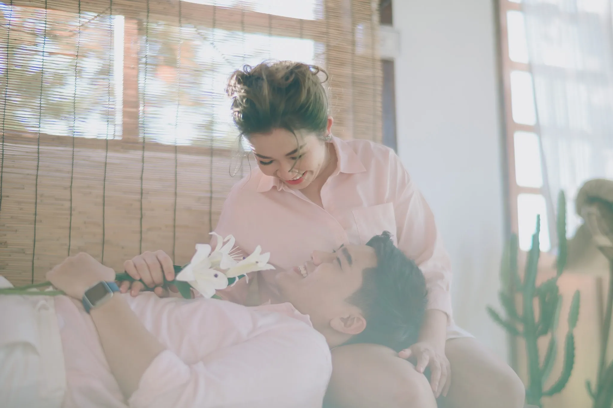 A laughing woman in pink leaning over a man lying in her lap holding white lilies