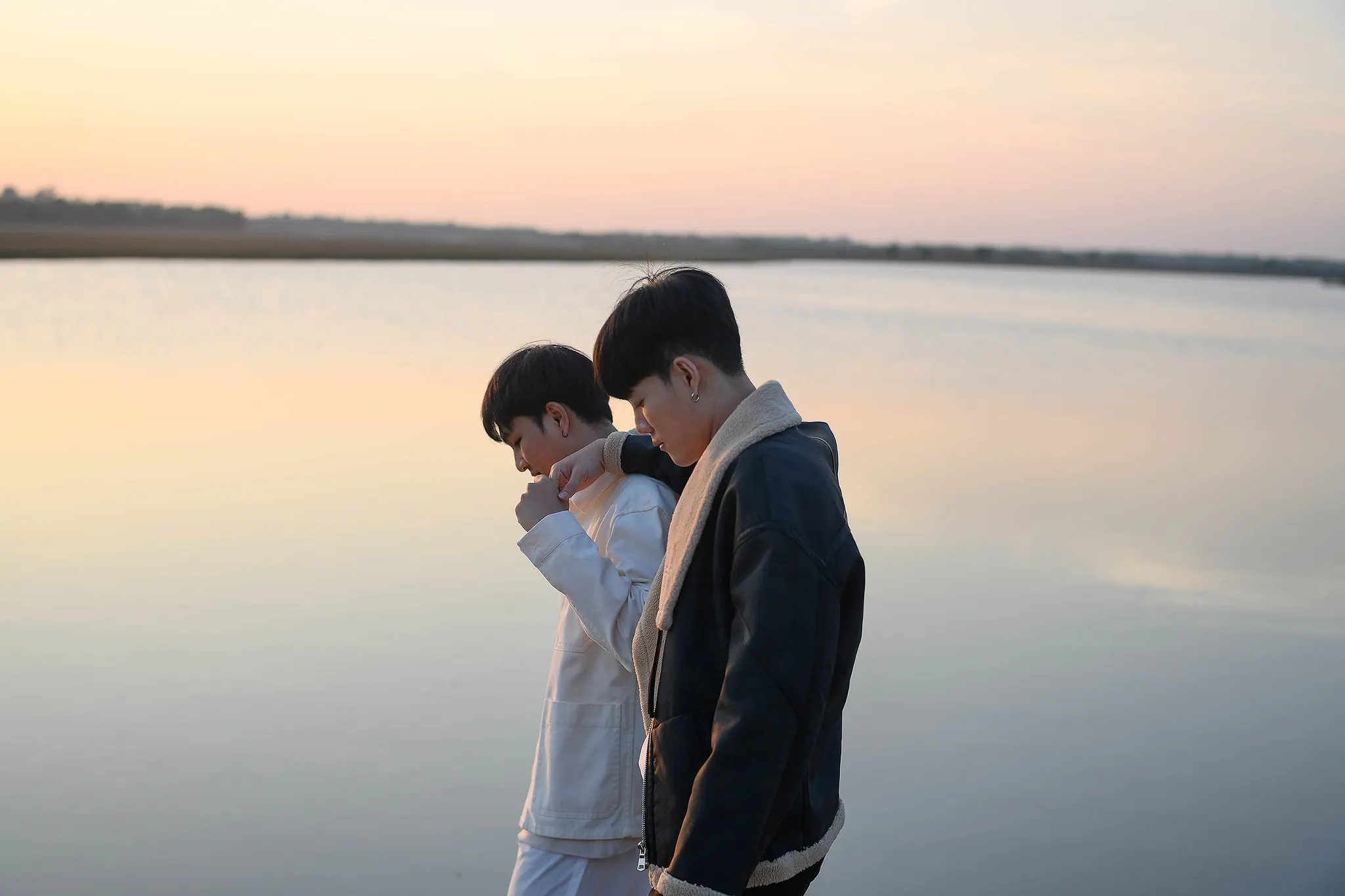 Two young men in light and dark jackets walking arm in arm beside a lake at dusk
