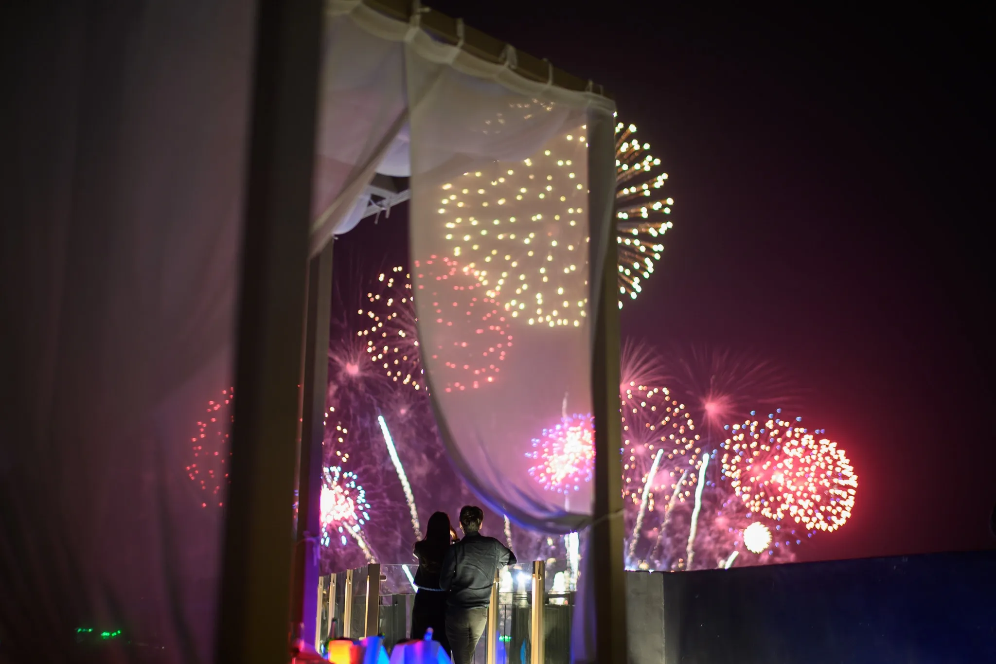 A couple watching fireworks from a curtained rooftop terrace at night