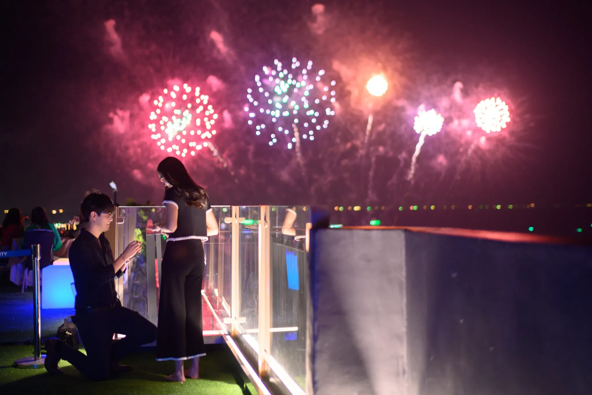 A man kneeling to propose on a rooftop terrace as fireworks burst over the water