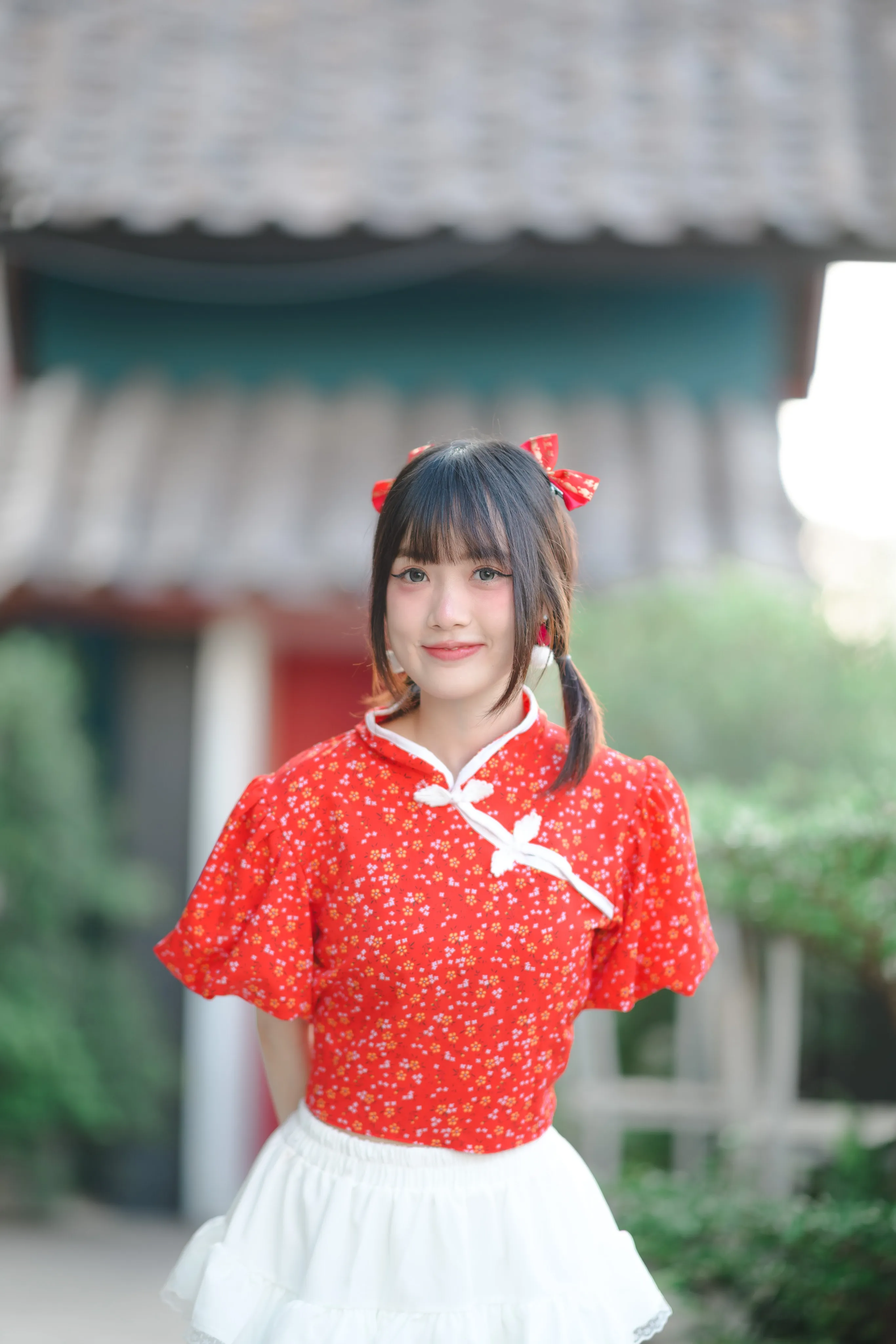 A smiling woman in a red floral top with red hair bows in front of a blurred wooden building