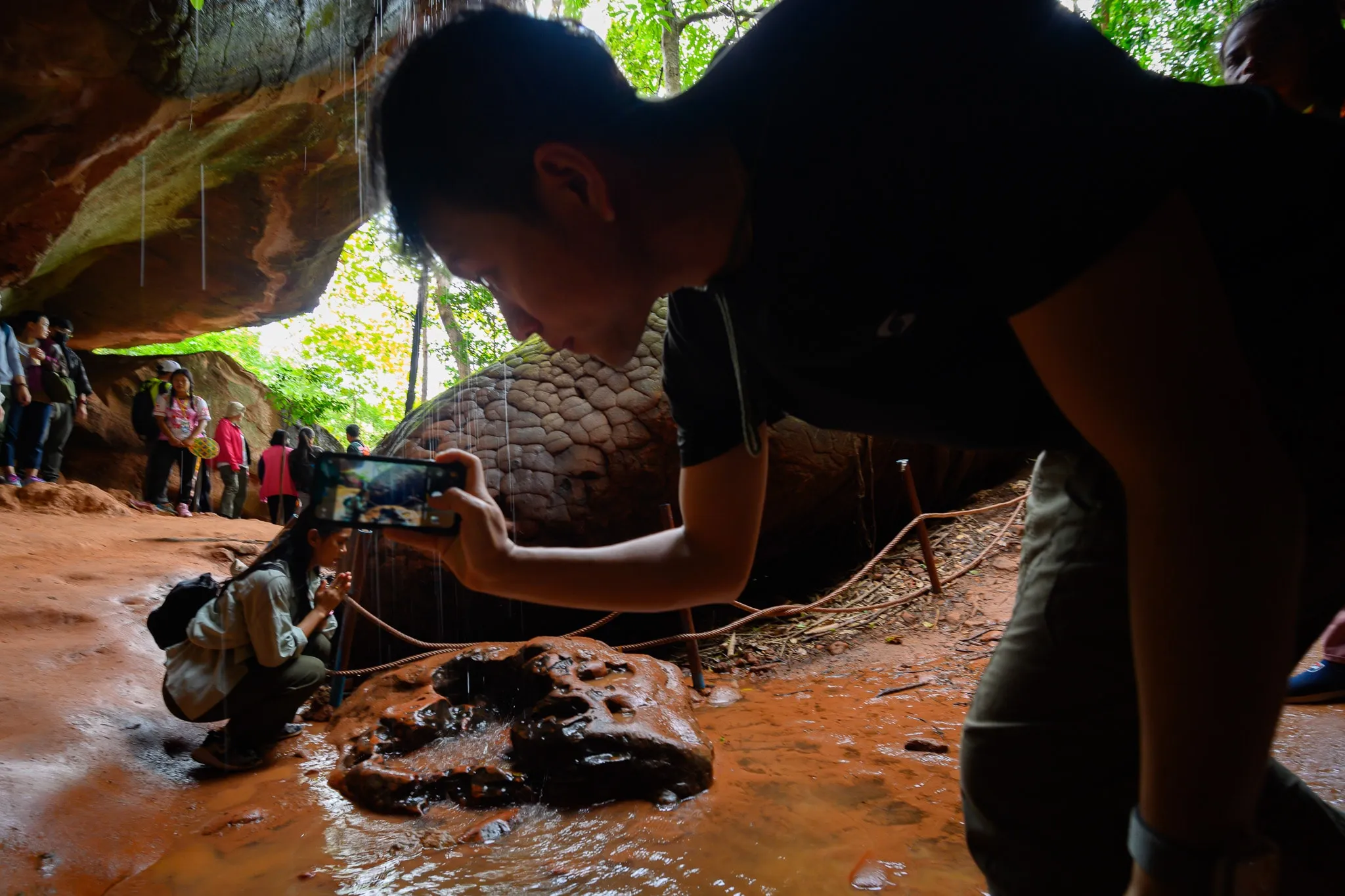 A visitor crouching to photograph a rock in a sandstone cave while tourists pass behind