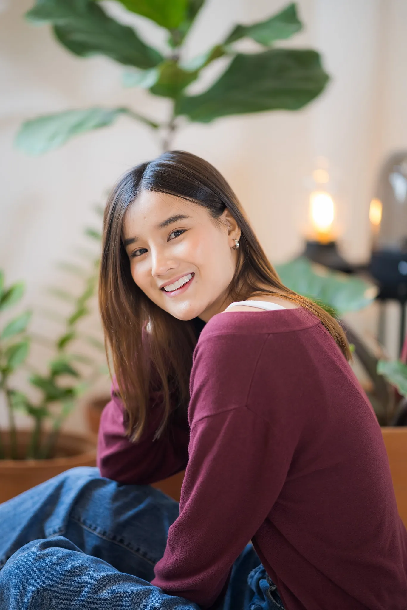 A smiling woman in a maroon sweater sitting beside a potted fiddle-leaf fig
