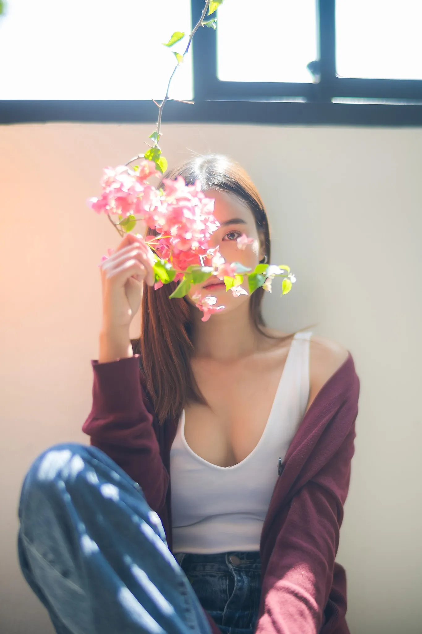 A woman holding a sprig of pink bougainvillea in front of her face in window light