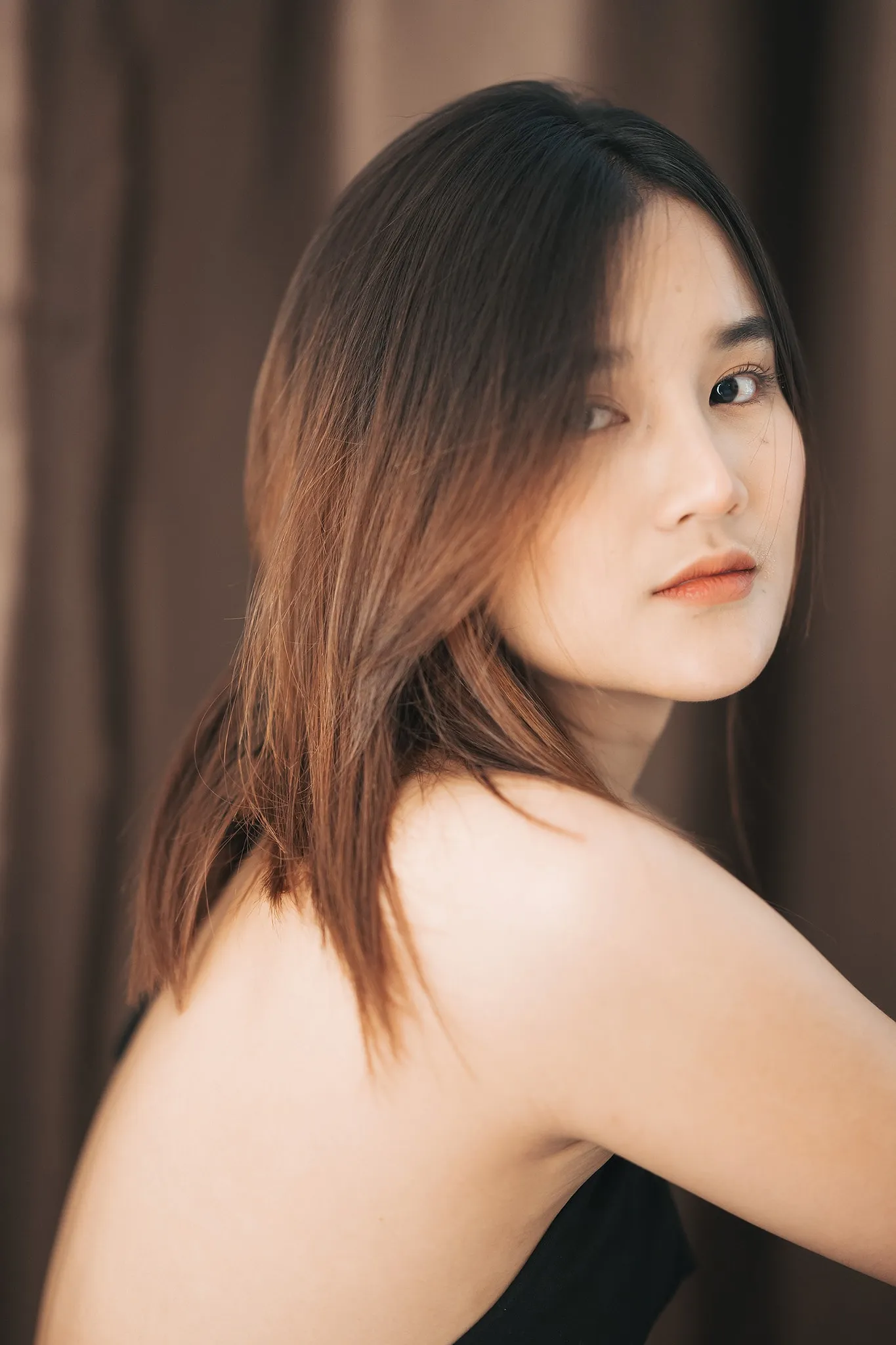 A woman with shoulder-length hair looking over her bare shoulder against a brown curtain