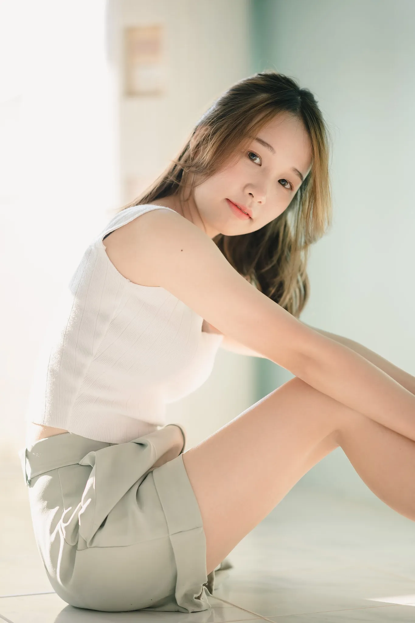 A woman in a white top and grey shorts sitting on a bright tiled floor, hugging her knees