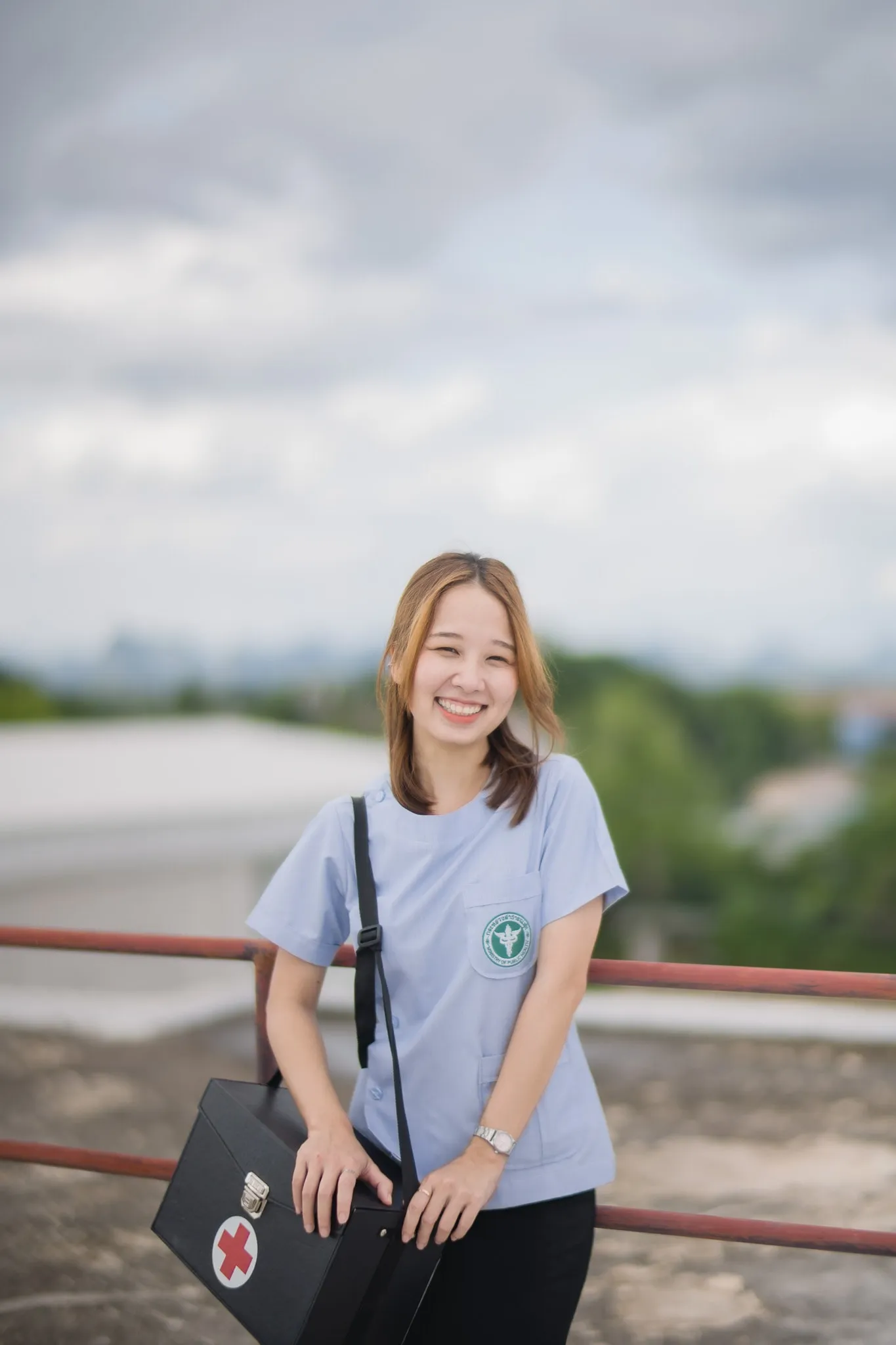 A smiling woman in a light blue uniform holding a first-aid case on a rooftop