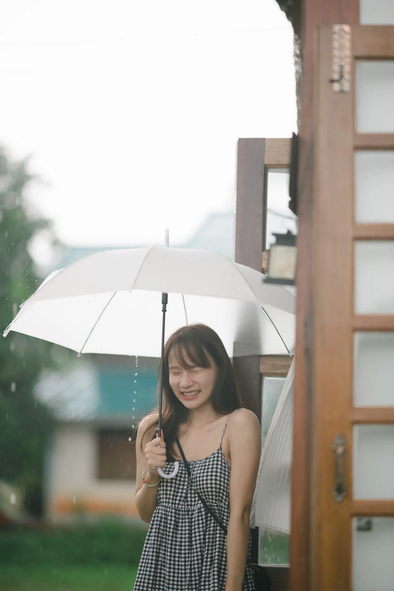A laughing woman in a gingham dress holding a white umbrella in the rain beside a wooden door