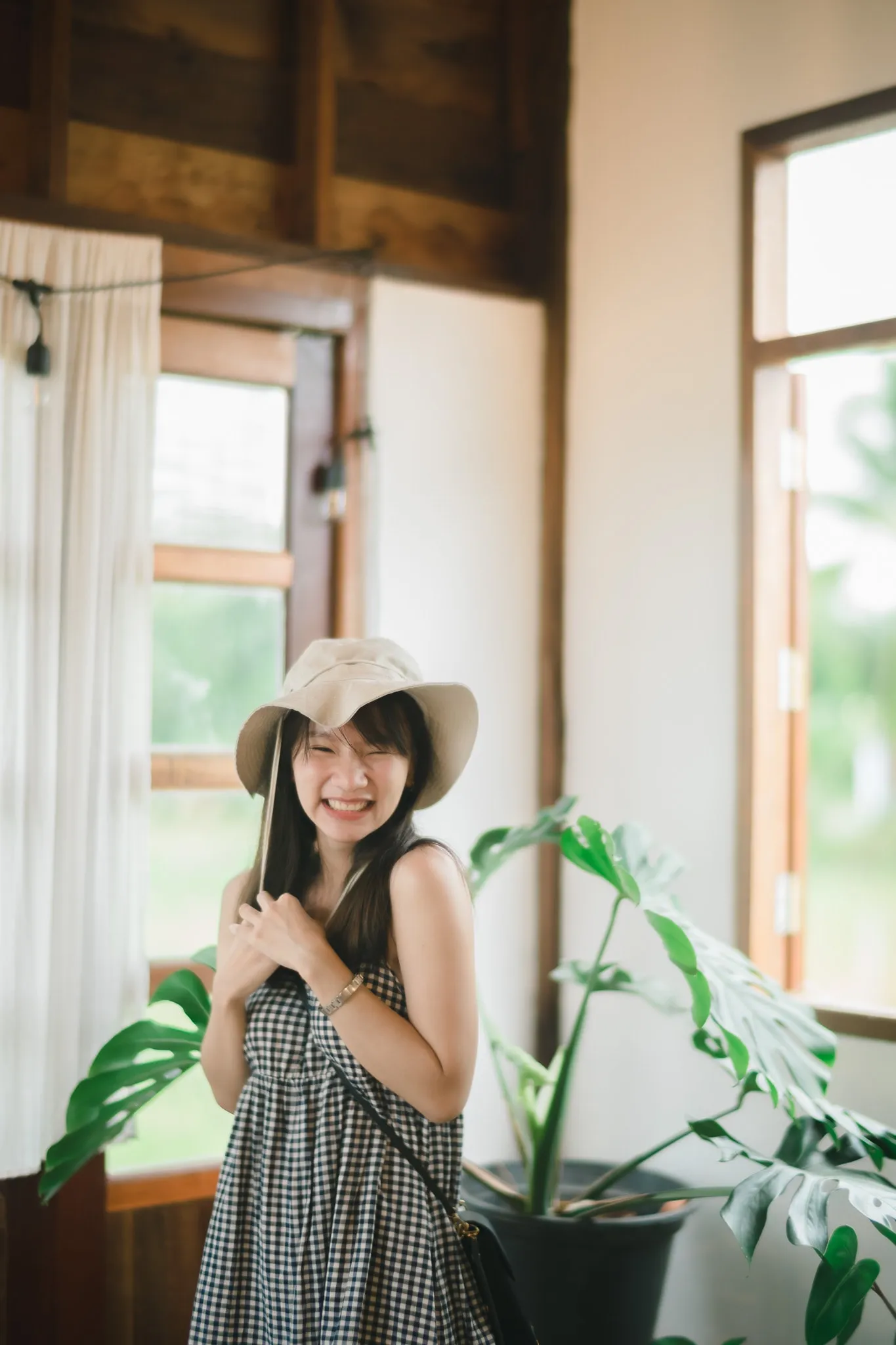 A smiling woman in a gingham dress and sun hat standing beside a monstera plant in a wooden house