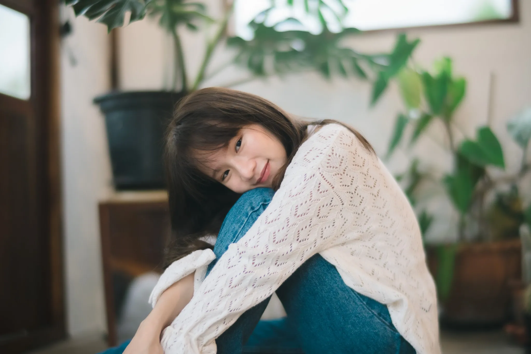 A woman in a white knit sweater and jeans hugging her knees among houseplants