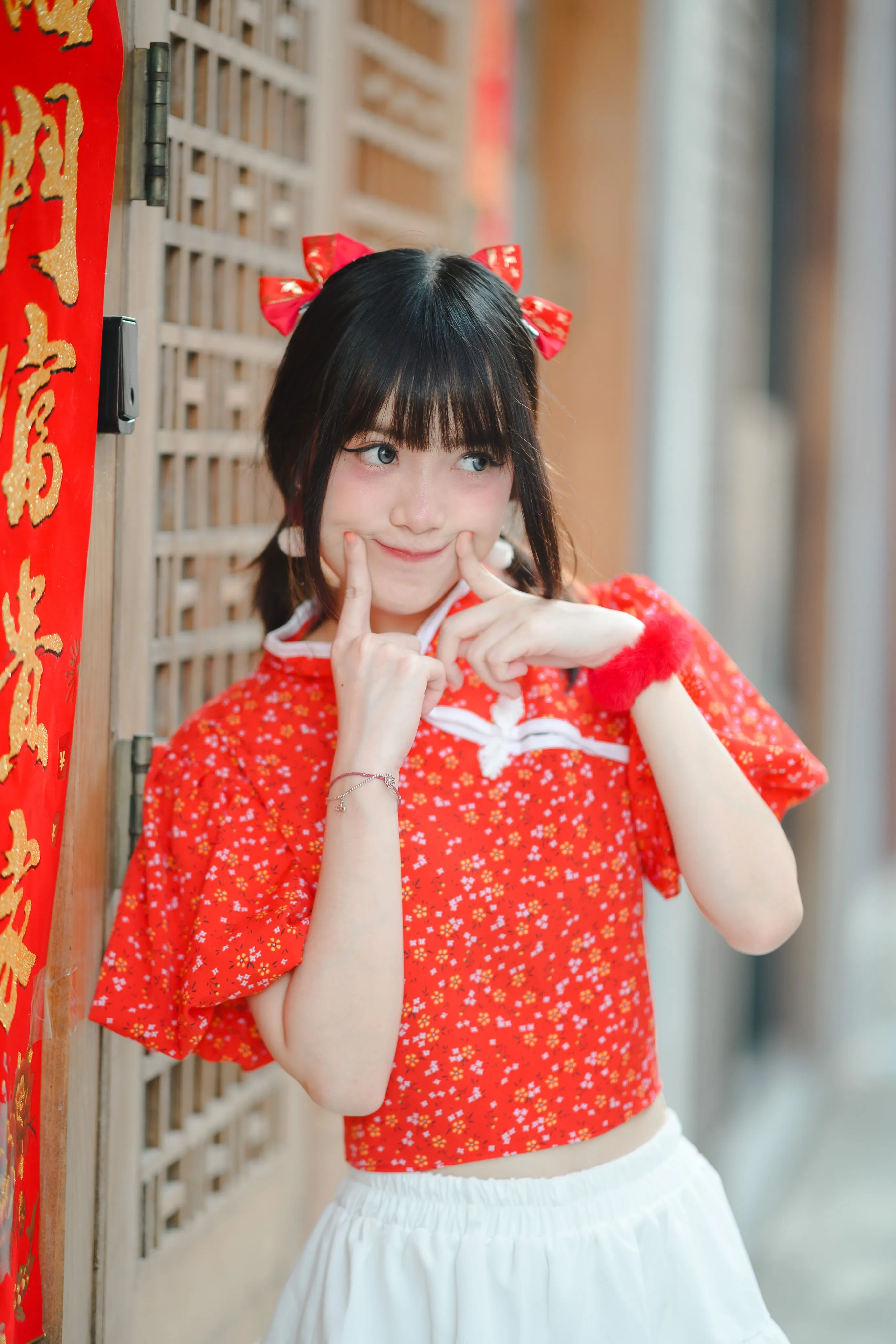 A woman in a red floral top and red hair bows framing her face with her fingers beside a red Chinese couplet