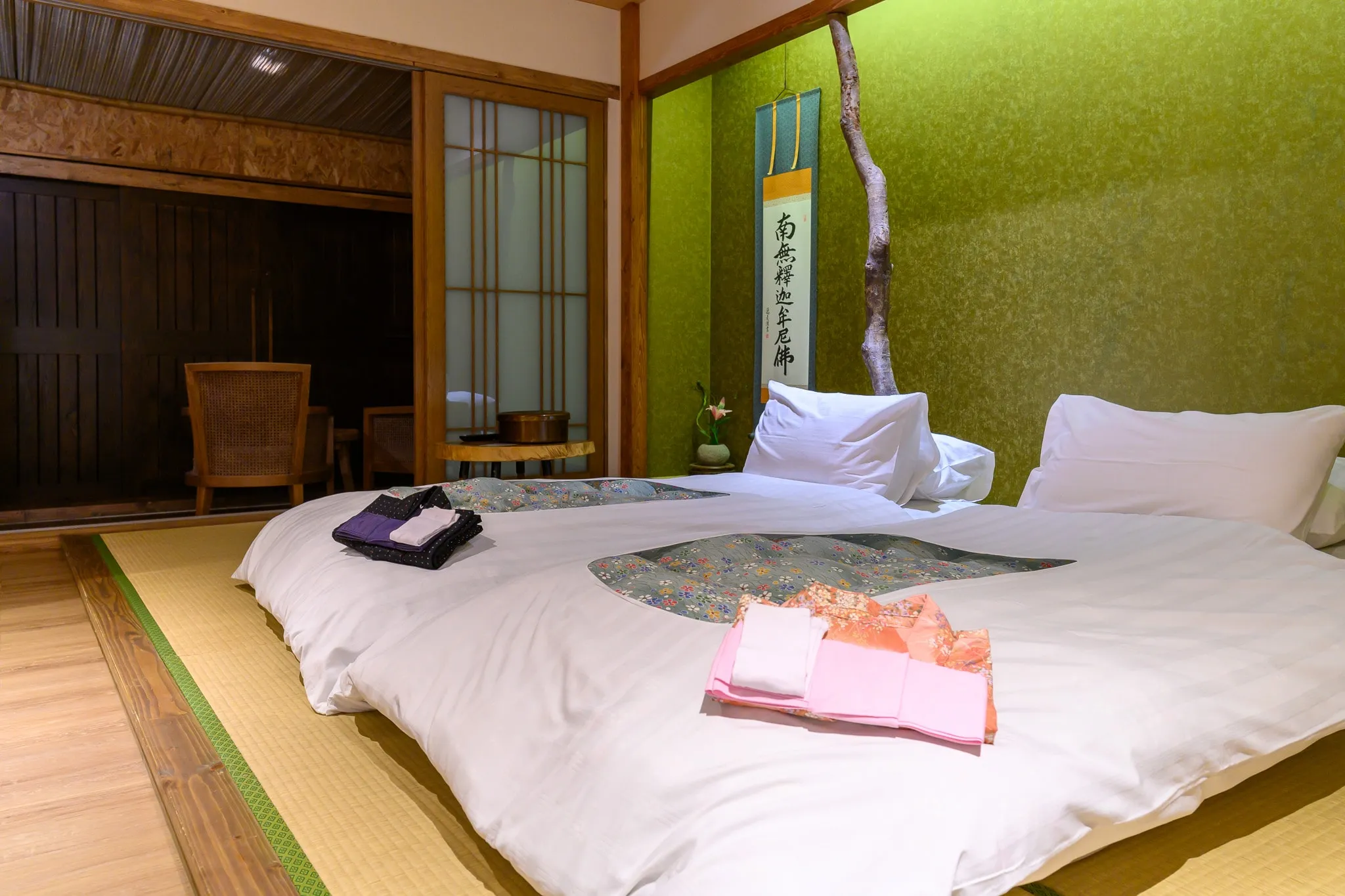 A Japanese-style guest room with a futon on tatami mats, folded yukata, and a hanging scroll